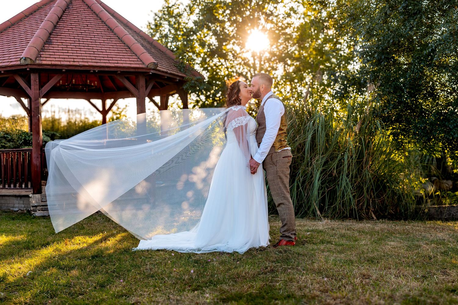 A flowing wedding dress catches the golden sunlight during a romantic sunset photoshoot by a wooden gazebo.