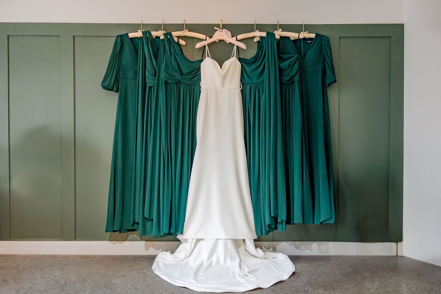 White wedding dress and emerald green bridesmaid dresses hanging on a wall displaying bridal party attire.