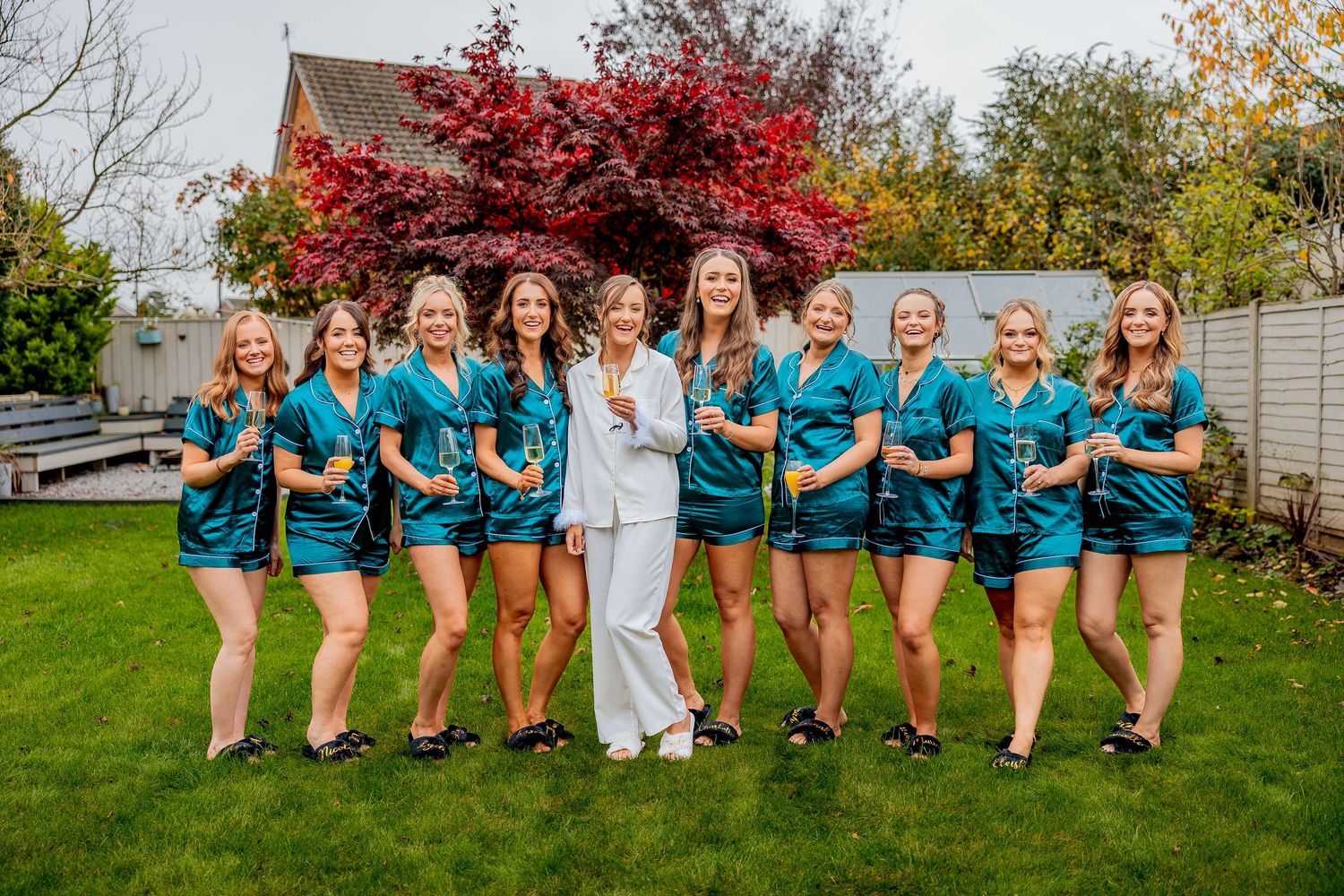 Group wearing matching teal bridal robes poses playfully in outdoor setting during fall season.