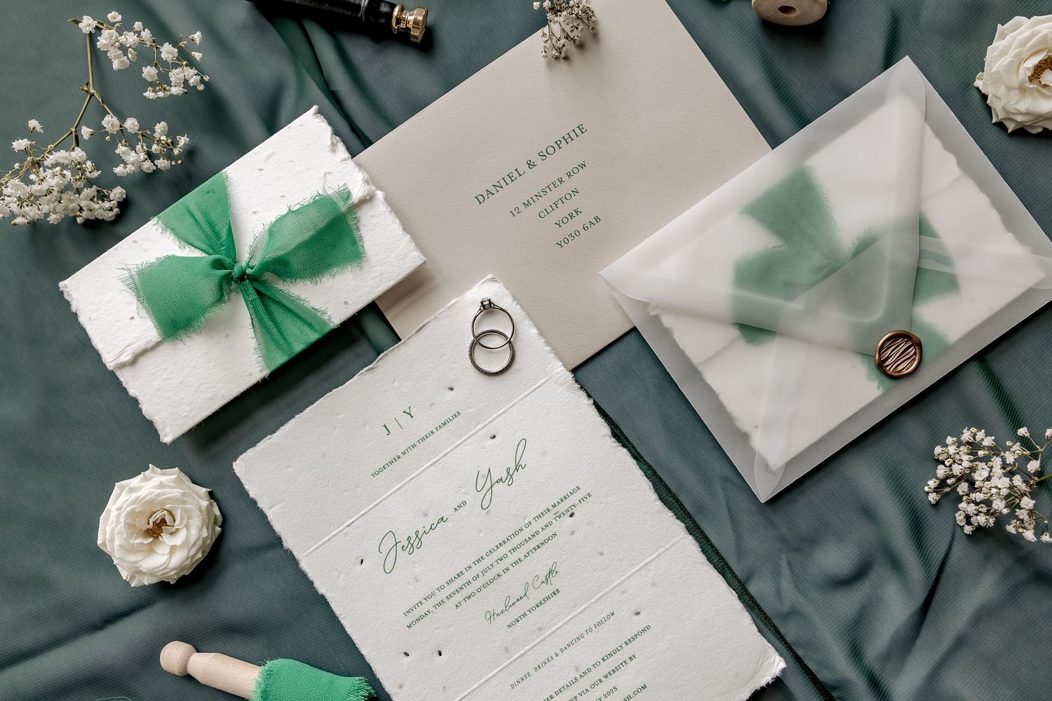 Elegant wedding invitations with green bows arranged on emerald silk fabric with white flowers and decorative elements.