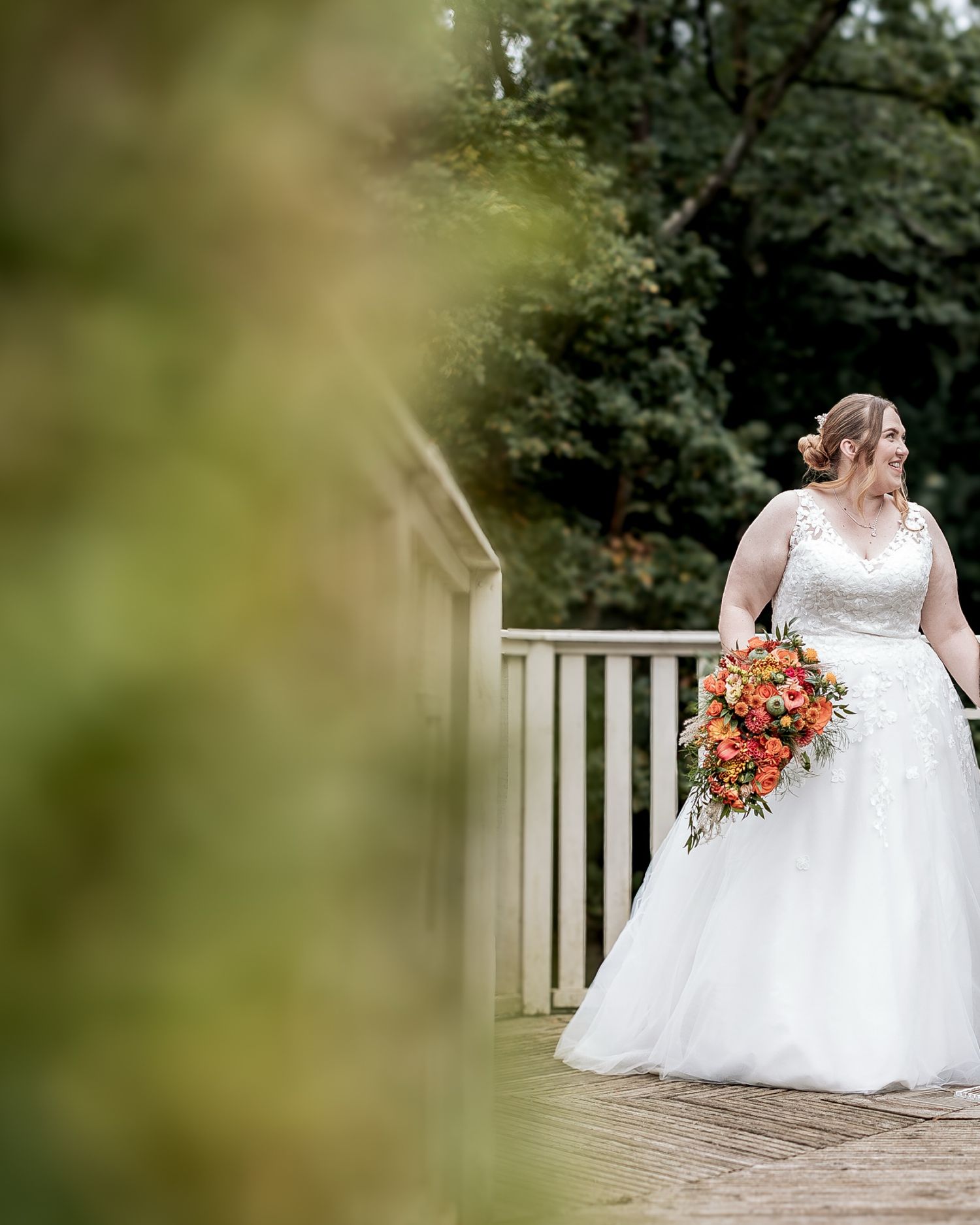 A bride in a white gown walks  on a wooden floor in a garden setting holding a vibrant orange and pink bouquet.