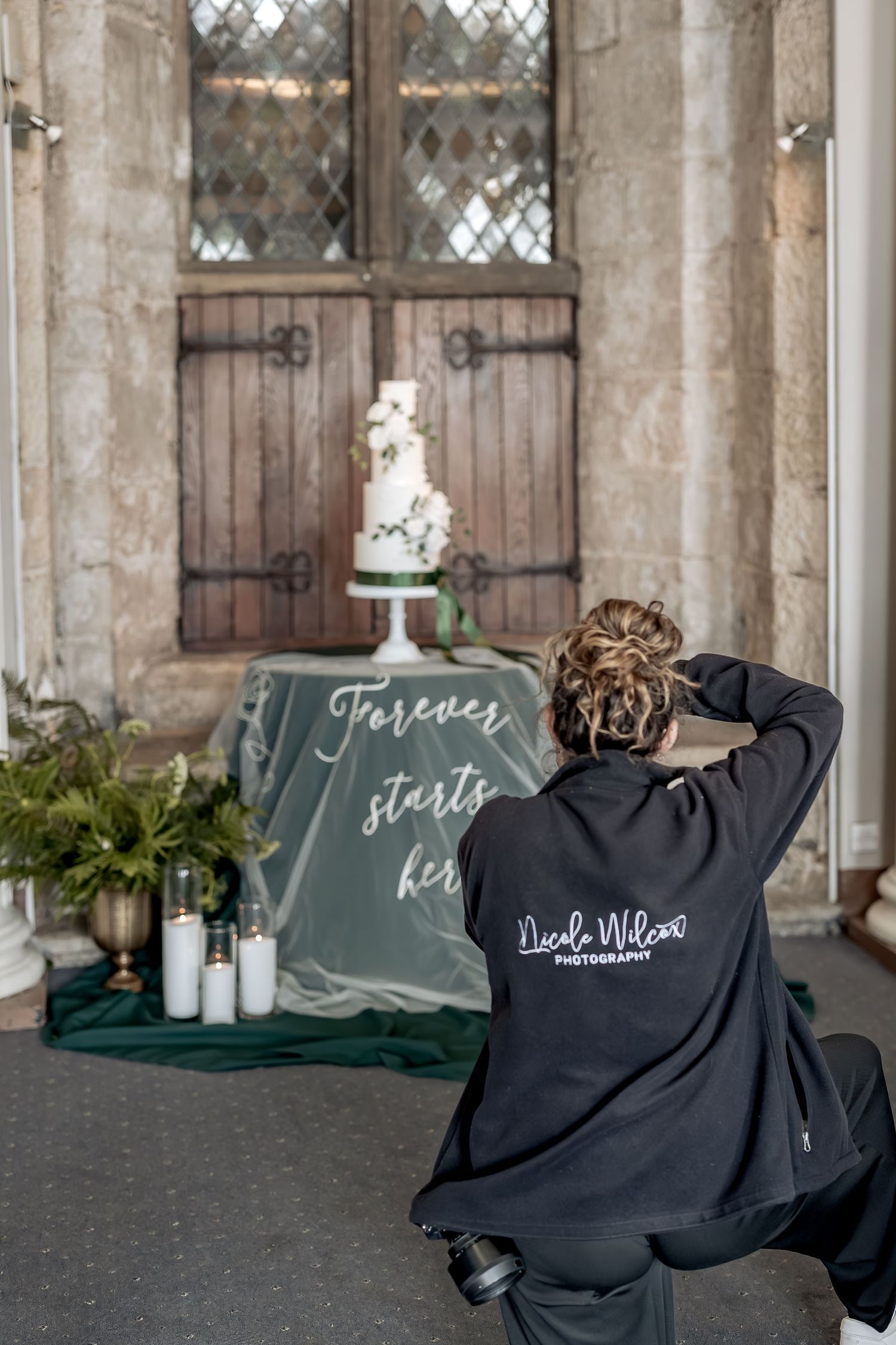 Wedding photographer captures a tiered white cake setup against rustic wooden doors in a historic stone chapel.