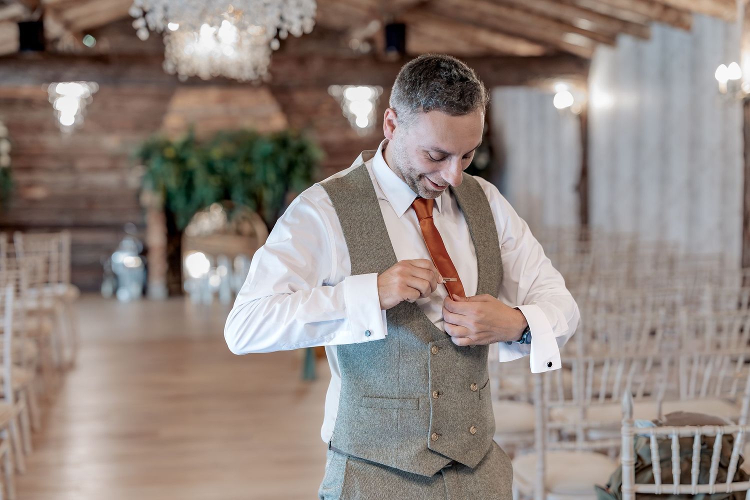 Groom in a gray vest and tie adjusts their outfit in a rustic wooden wedding venue with chiavari chairs.
