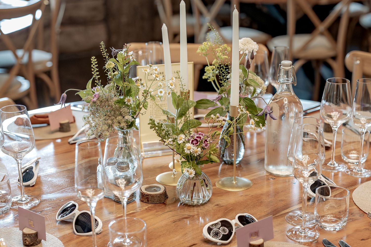 Elegant rustic table setting with white floral centerpieces, glass candle holders, and wine glasses at a wedding reception.