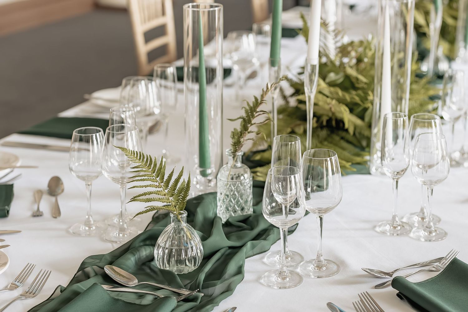 Elegant table setting with green tablecloth, wine glasses and tropical fern leaves in glass vases.