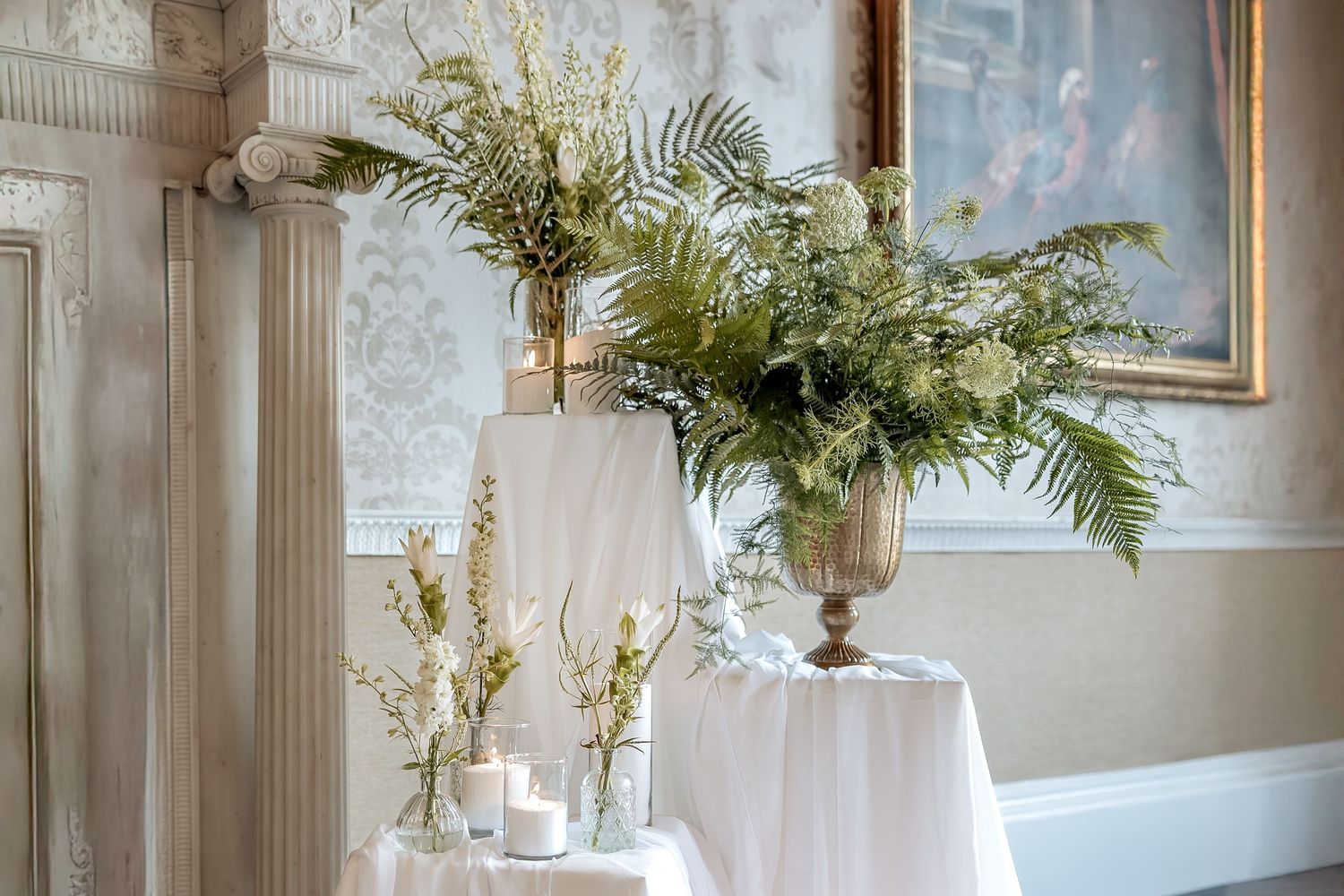 Elegant wedding decor features fern arrangements in silver urns on white-draped pedestals against damask wallpaper.