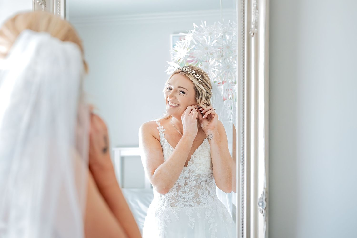 A bride in an elegant white lace wedding gown stands by a mirror during the getting ready process.