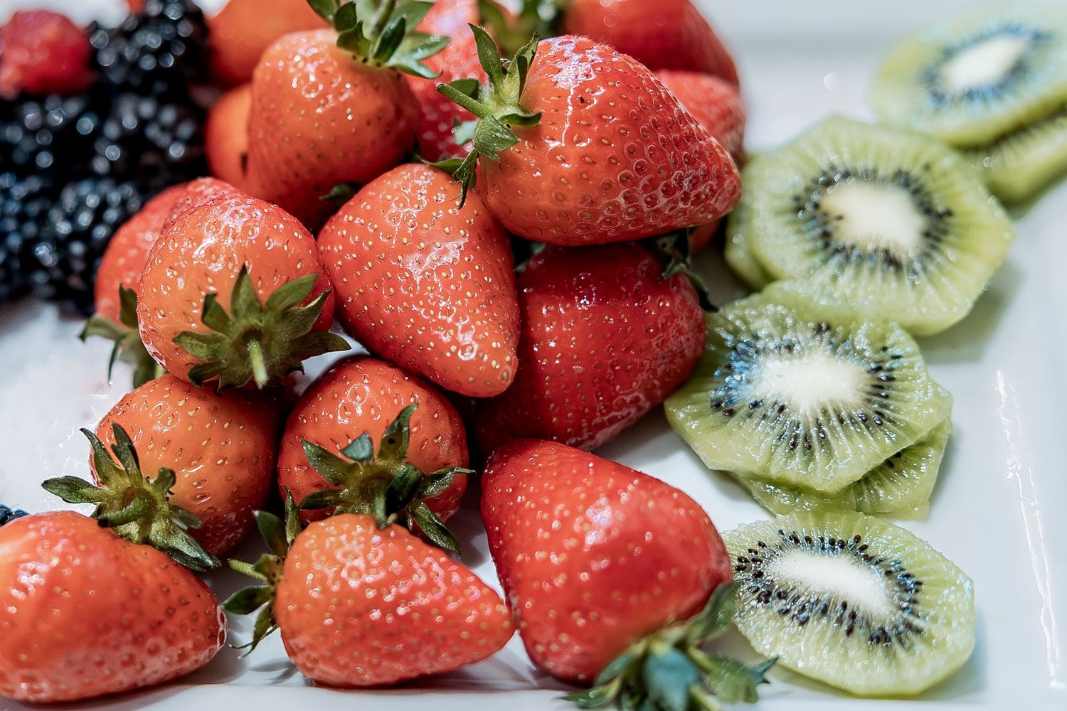 Fresh strawberries, blackberries and sliced kiwi fruit arranged on a white plate with water droplets.