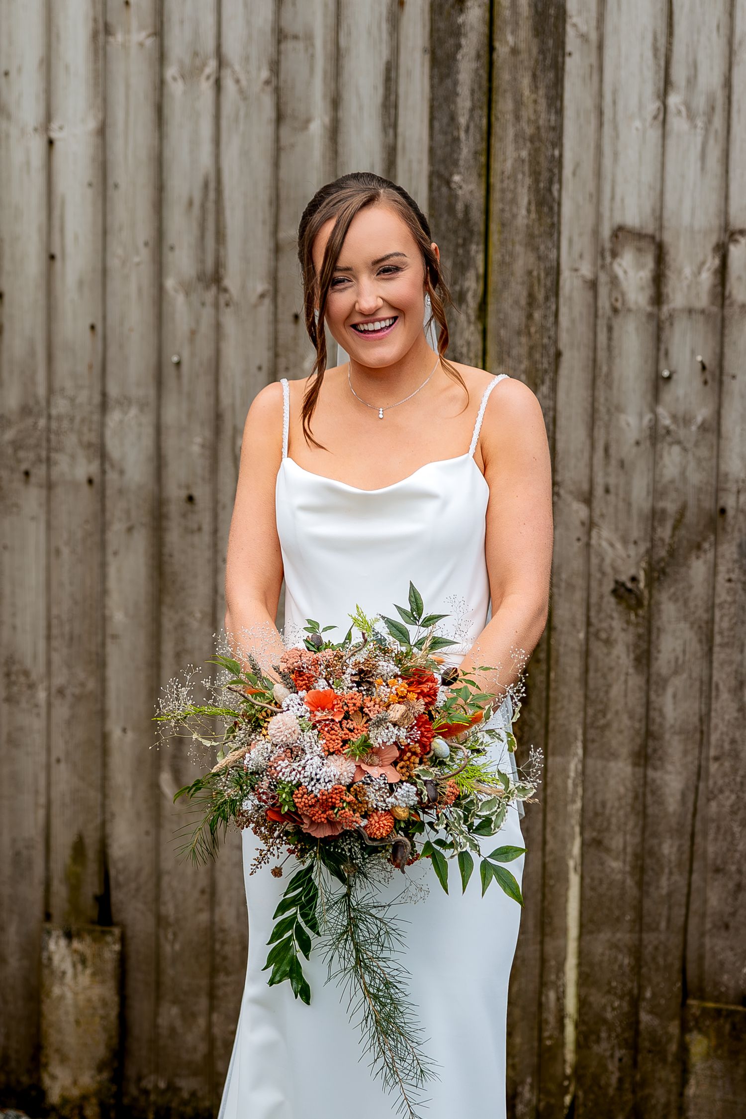 A bride in a white dress holds a vibrant orange and green floral bouquet against a rustic wooden fence backdrop.