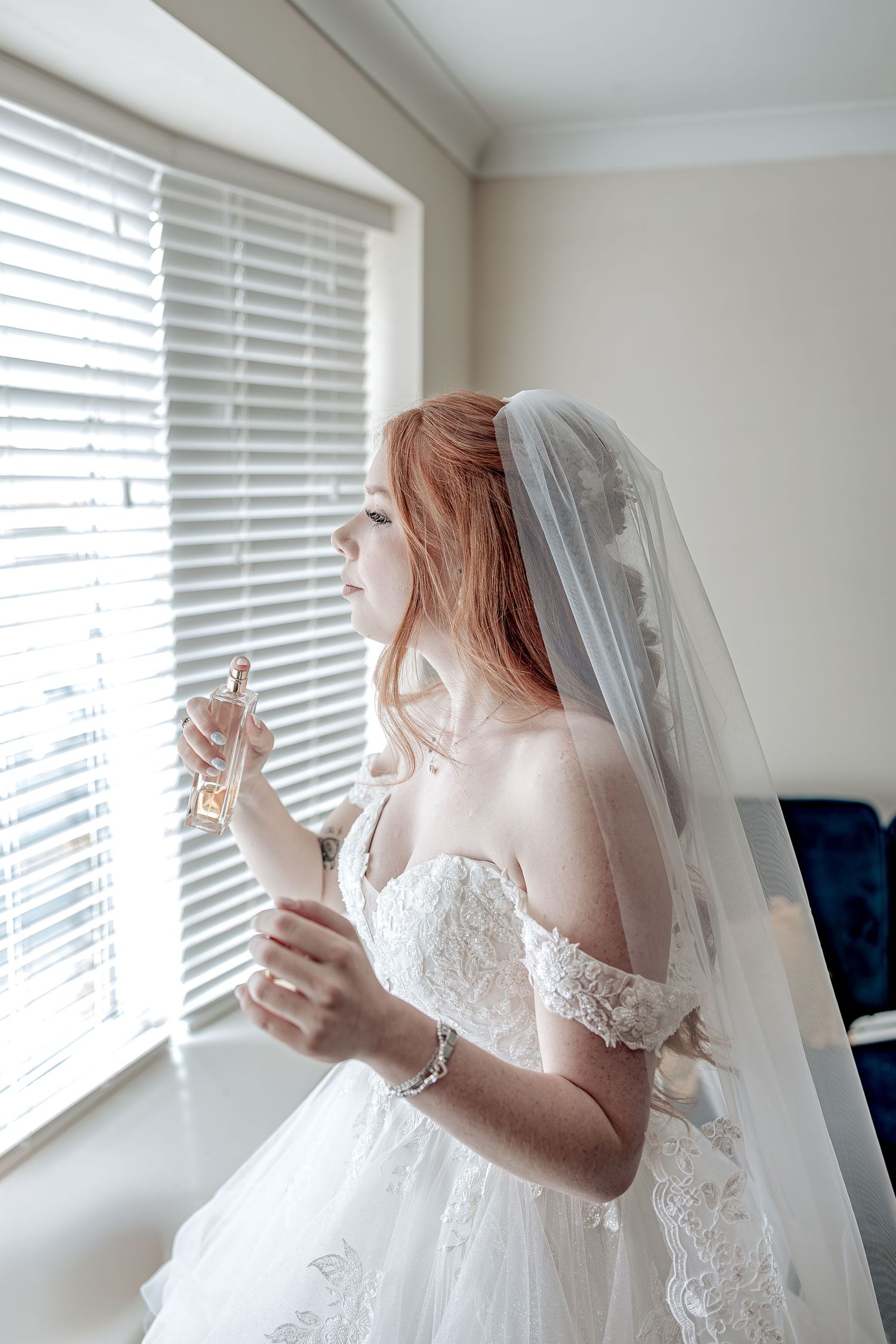 Bride in lace white wedding dress and cathedral veil poses by sunlit window blinds in a serene bridal portrait.