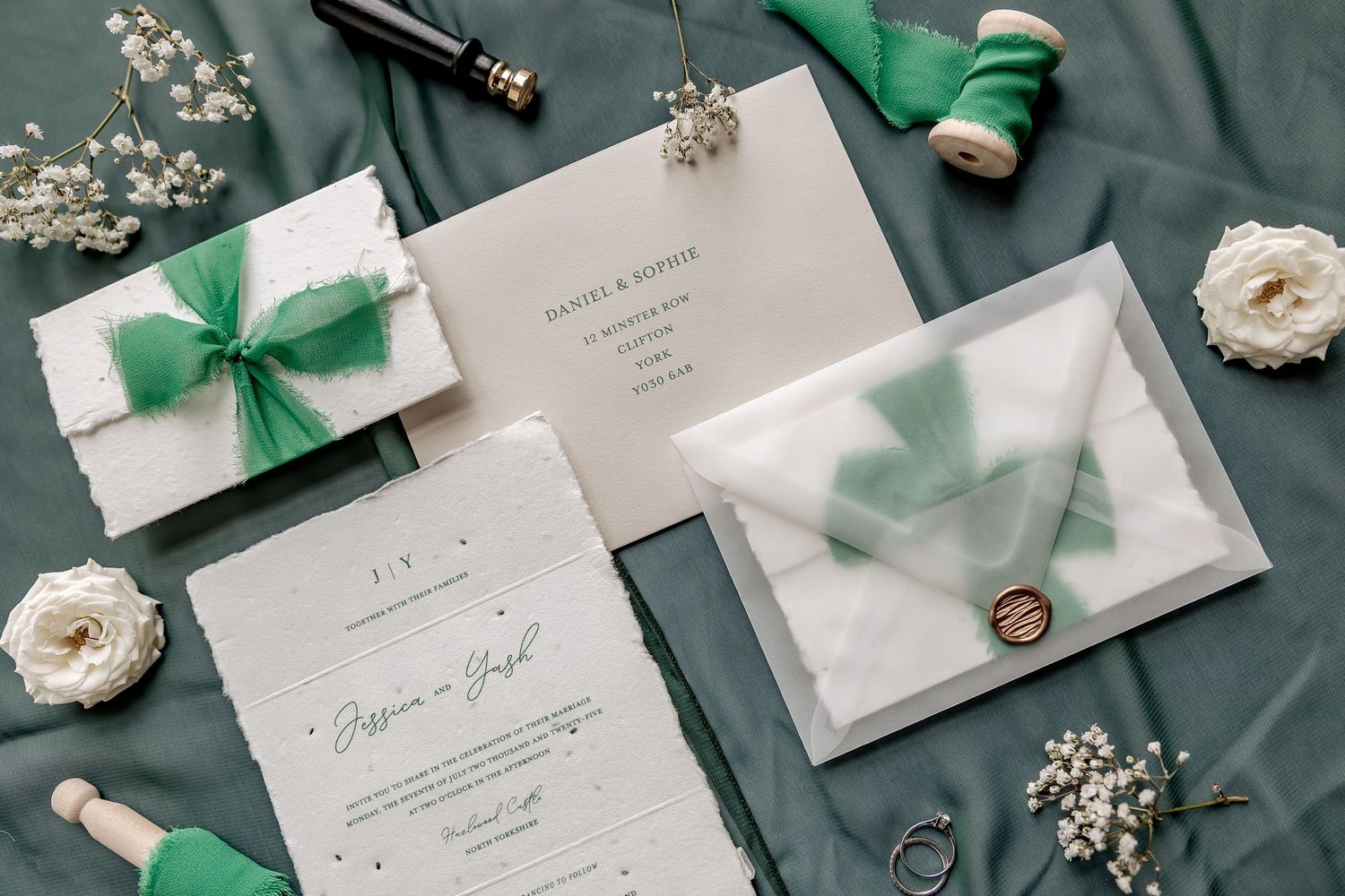 Elegant wedding invitation suite featuring white envelopes with mint green ribbons and delicate floral accents on green fabric.