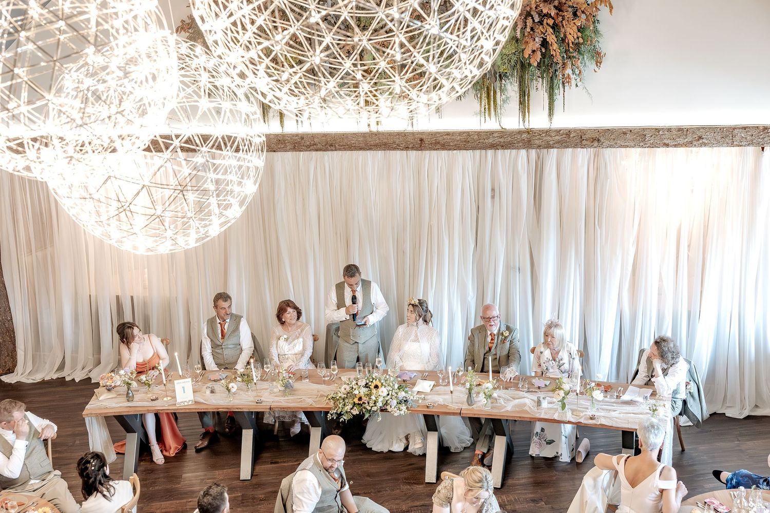 Elegant white wedding reception venue with crystal chandeliers and draped fabric decorating the ceiling and walls, during a speech.