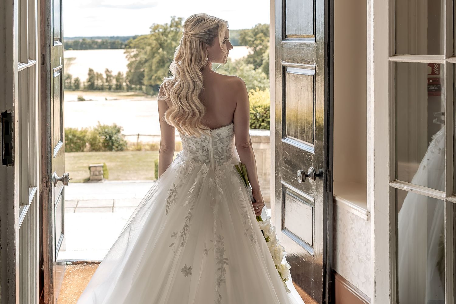 A bride in an embellished white ballgown stands in a doorway overlooking a scenic  view.