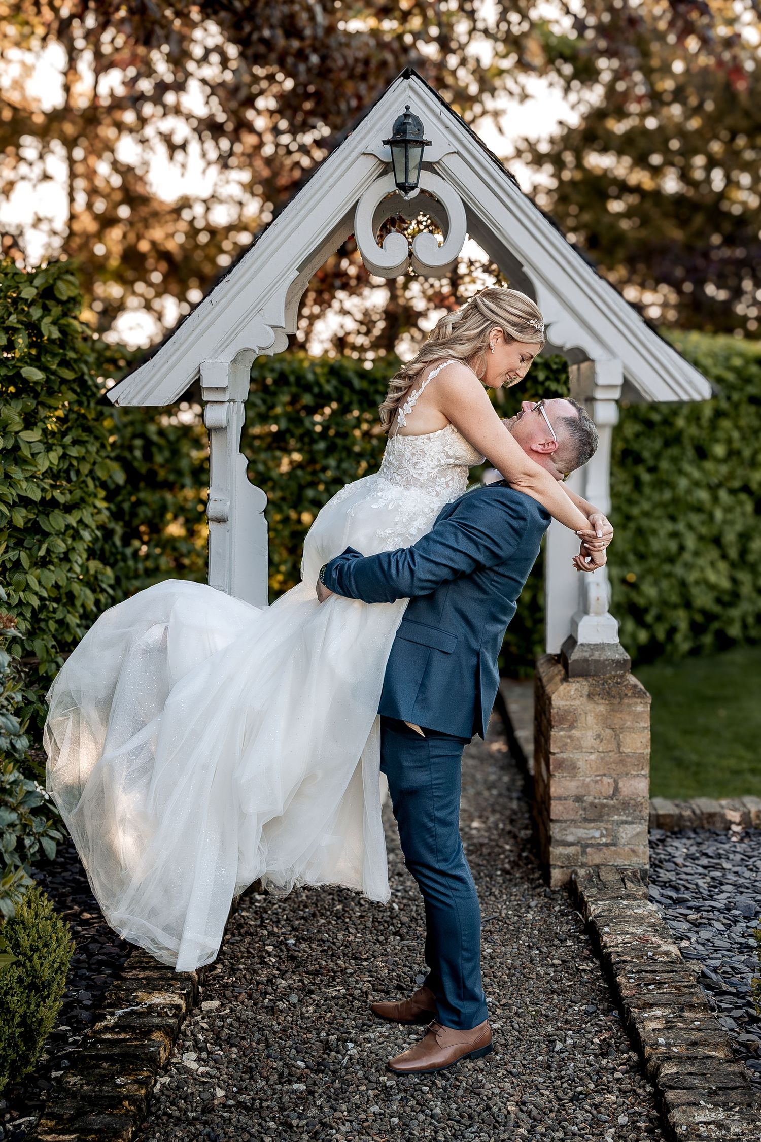 A romantic wedding couple dips and embraces in front of a white garden arbor at sunset.