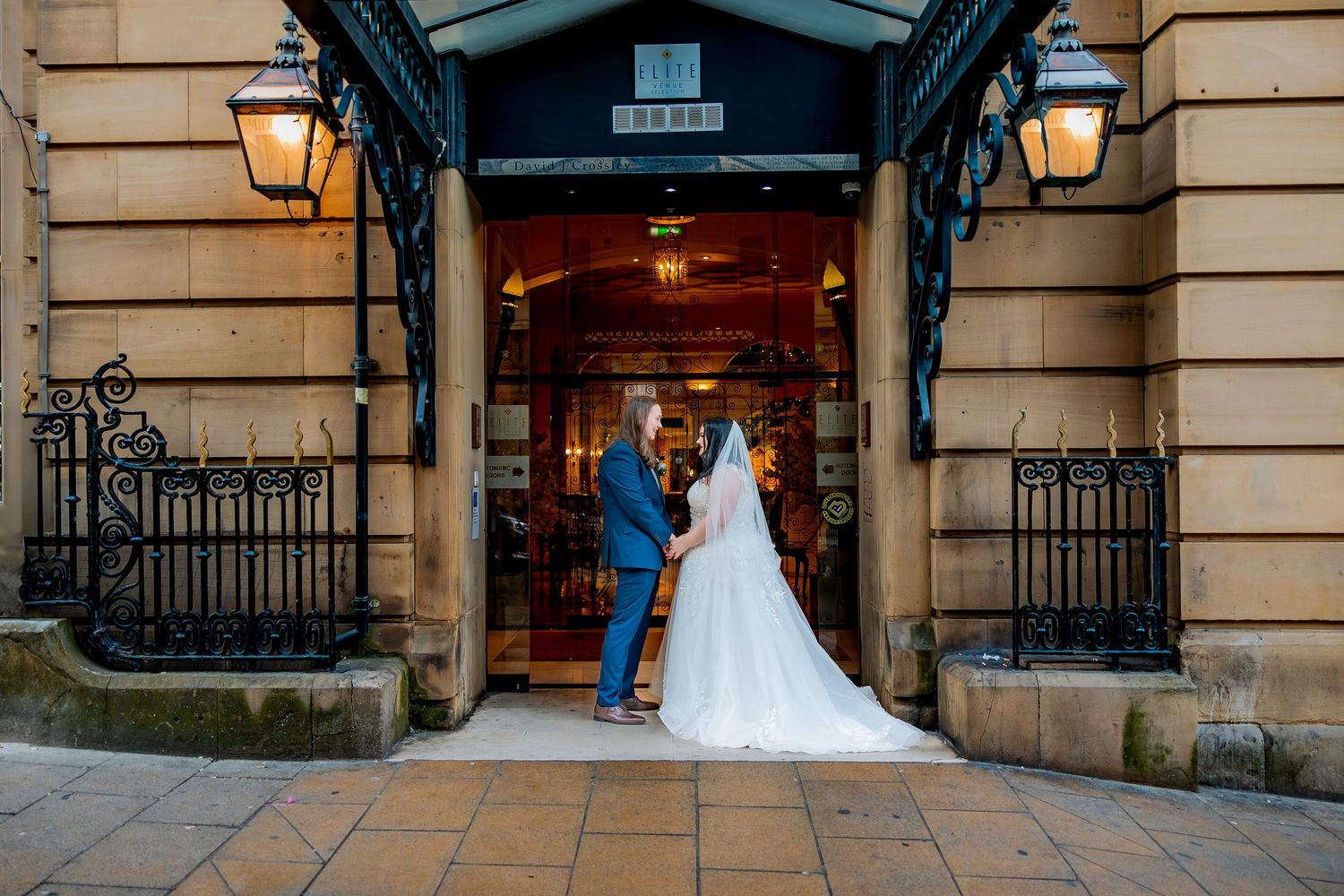 LGBTQ wedding couple poses in front of historic stone hotel with ornate lanterns and architecture.