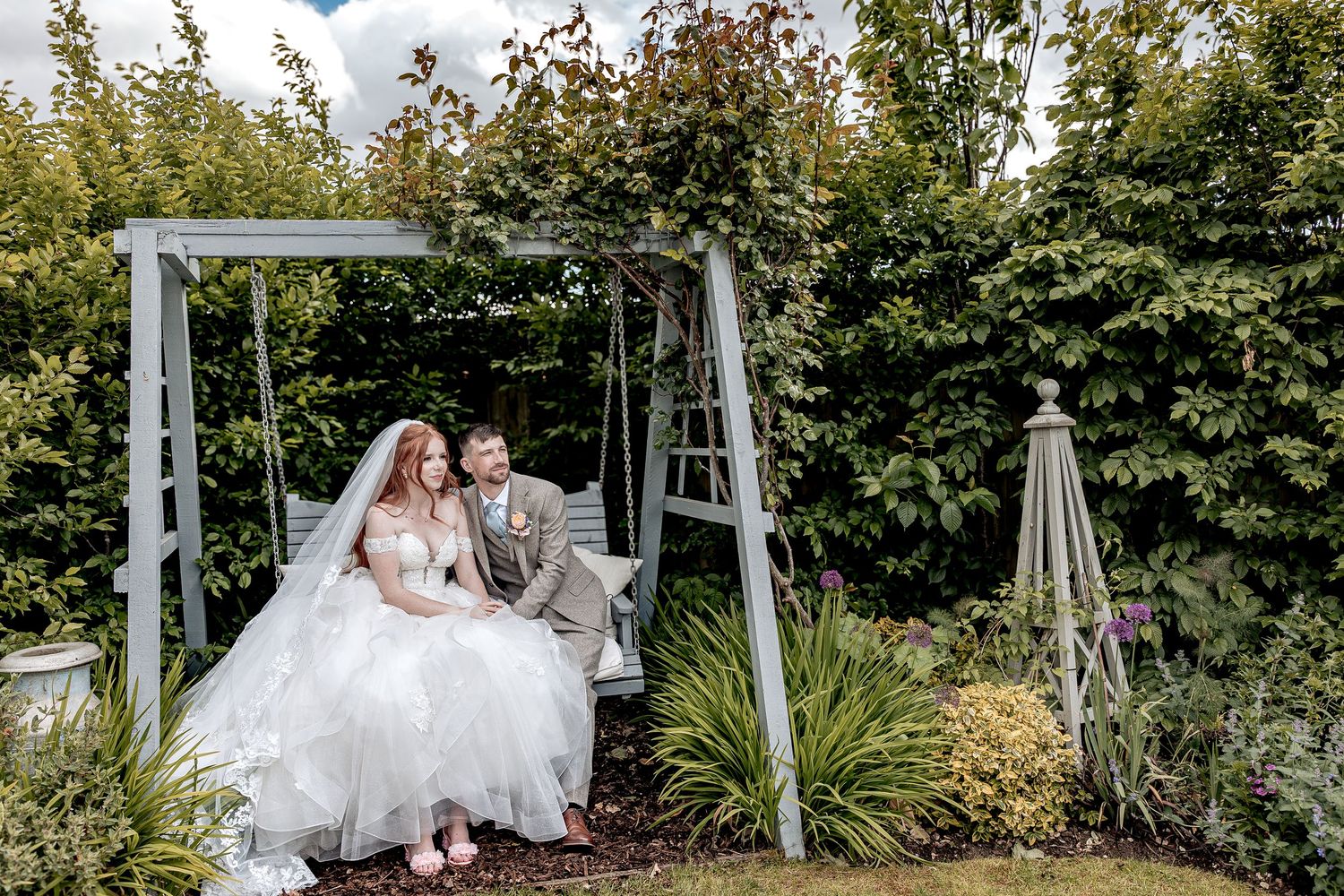 A romantic wedding couple sits together on a garden swing set surrounded by lush greenery on a sunny day.