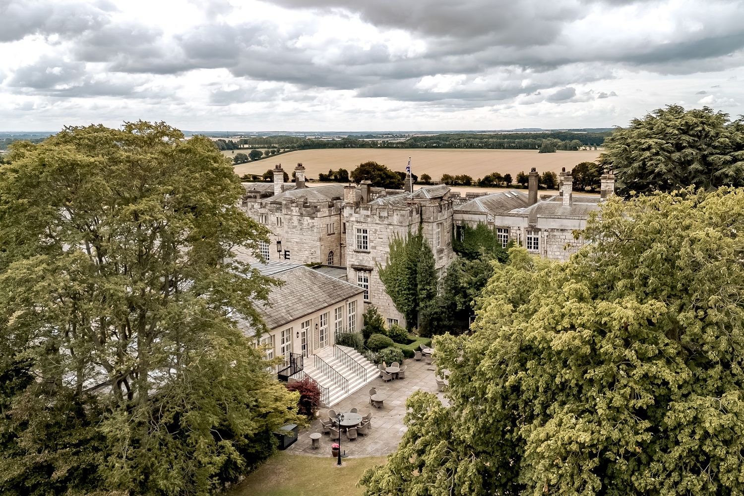 Elevated view of historic stone buildings in an English town with dramatic cloudy sky and green trees framing the scene.