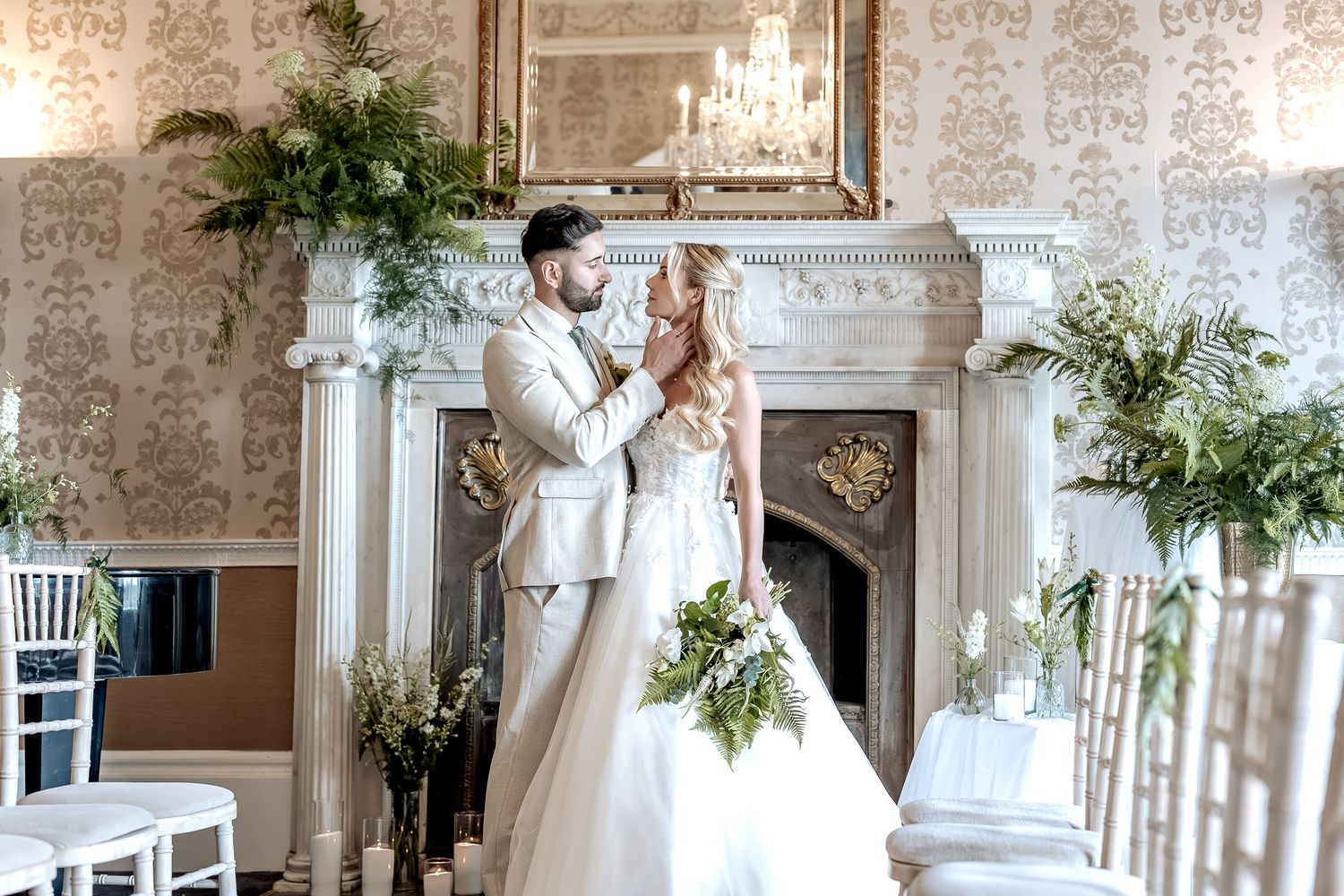 Elegant white wedding venue interior with ornate columns, vintage wallpaper, and cascading greenery decorations throughout.
