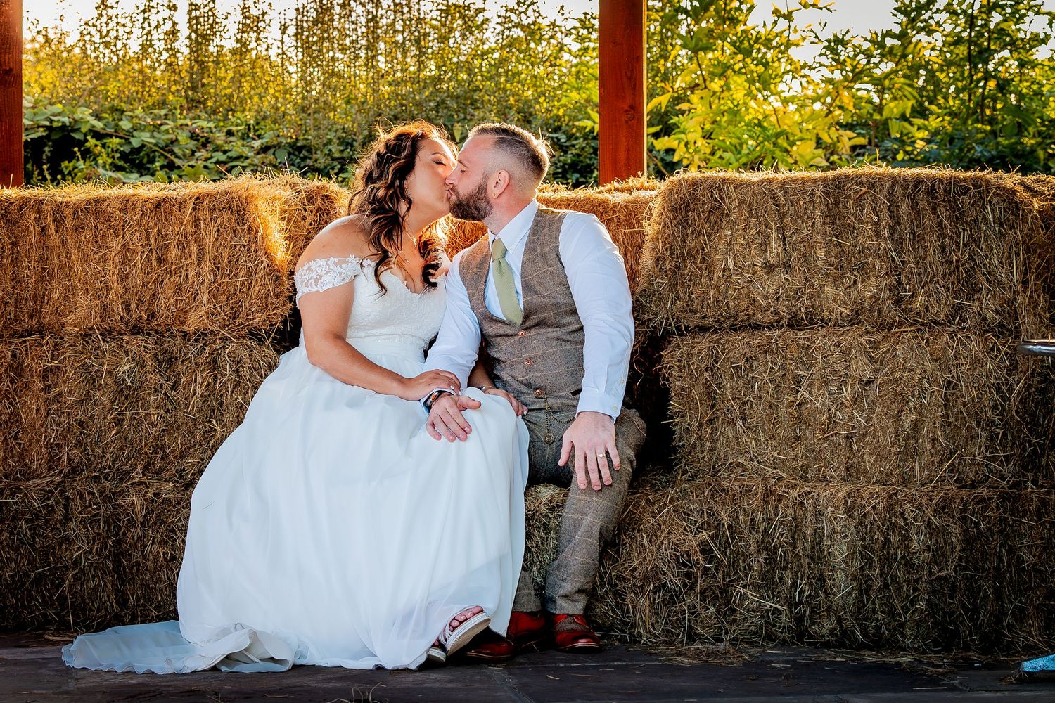 A romantic couple in wedding attire sits together on hay bales at sunset.