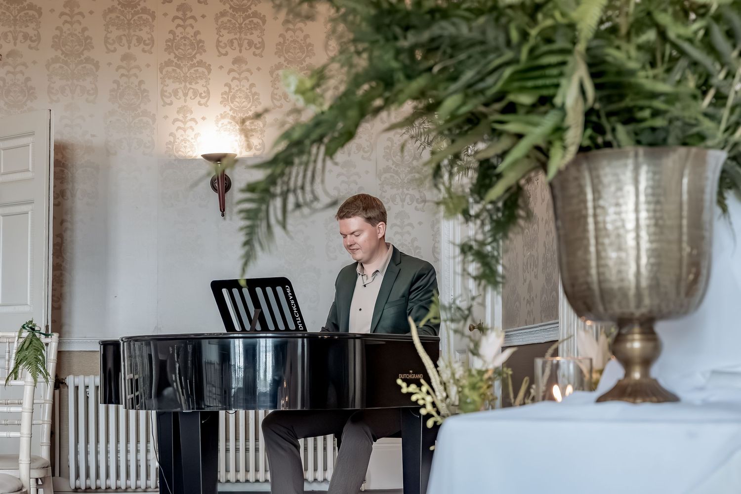 A pianist in a green jacket plays at an elegant venue with hanging plants and white brick walls.