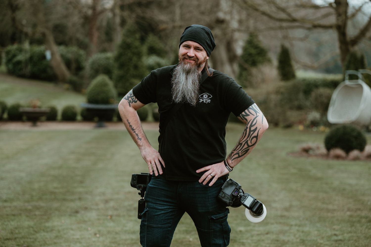 Photographer wearing a black t-shirt and beanie stands in a garden setting with camera gear on belt.