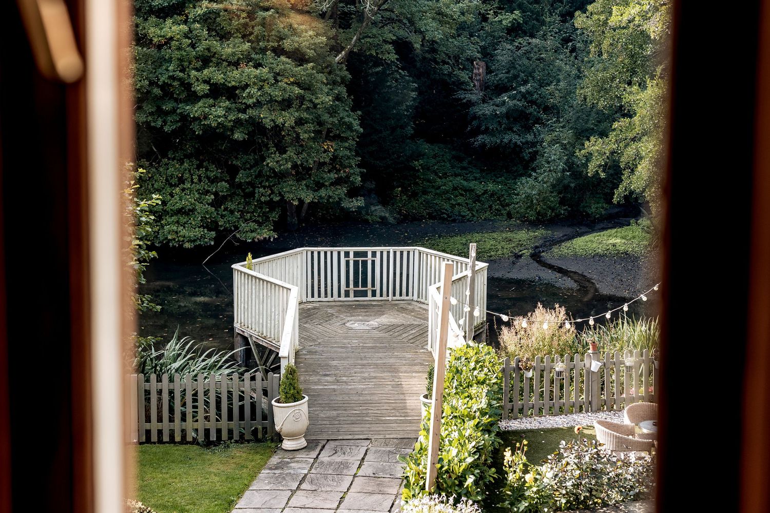 A white wooden gazebo with railing sits at the end of a stone path surrounded by lush garden landscaping and trees.