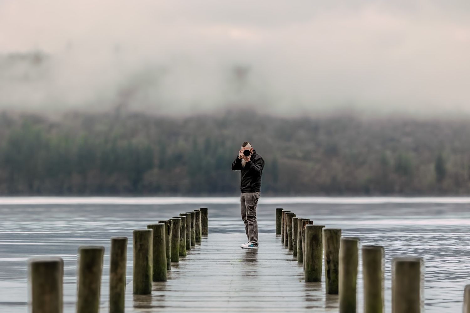 A photographer stands on a wooden pier extending into a misty lake in the early morning light.