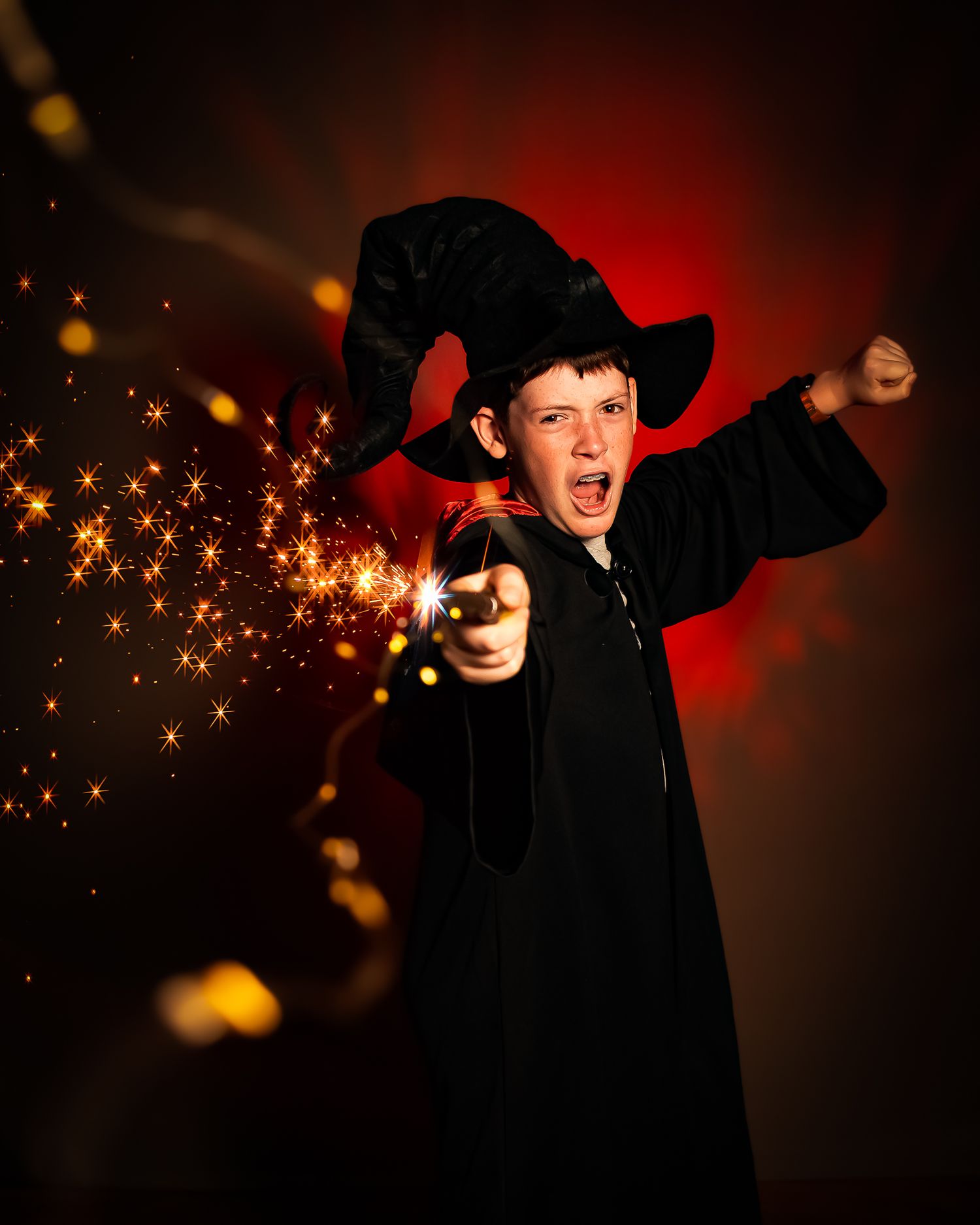 A boy in a black wizard costume and hat casting a spell with a magic wand emitting golden sparks.