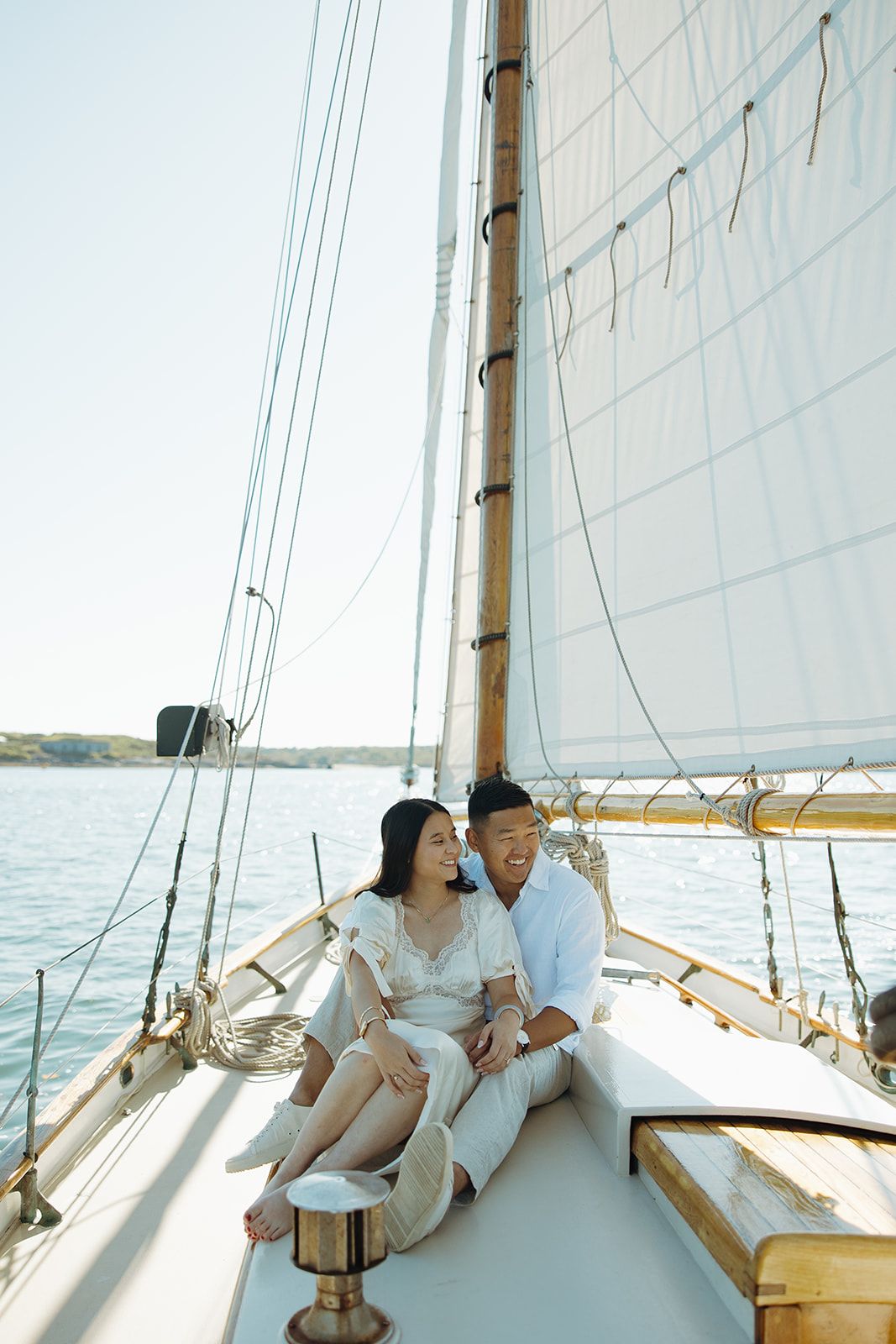 Couple relaxing on a sailboat deck in white attire against white sails on a sunny day at sea.