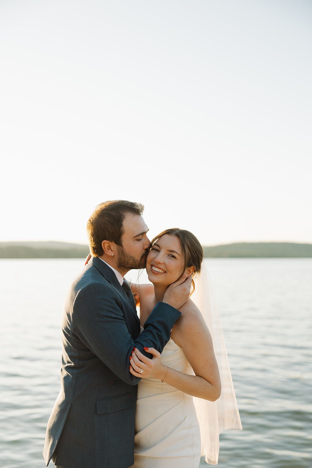 A romantic couple embraces and kisses by the ocean at sunset with water and distant hills in the background.