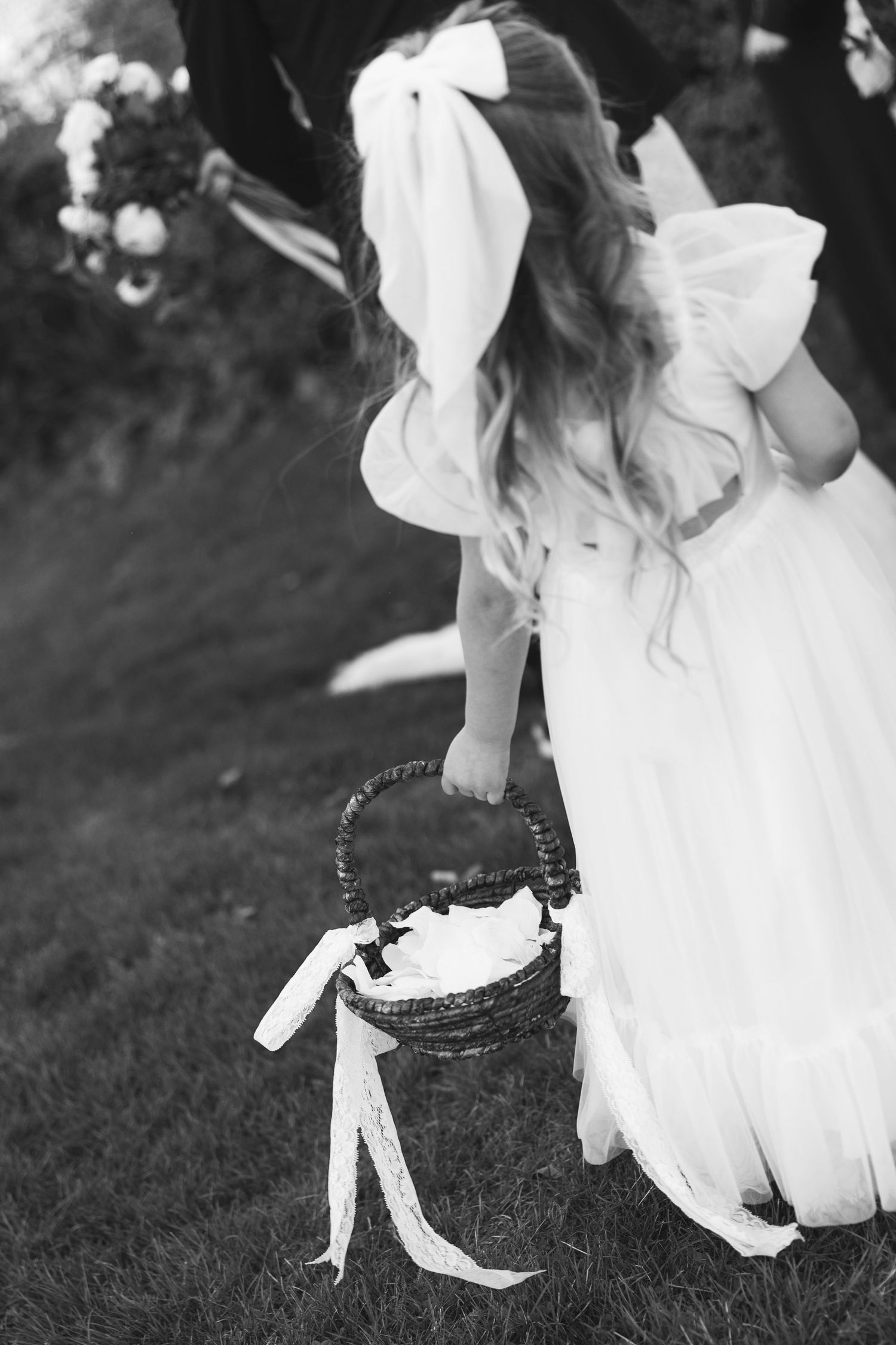 A black and white photo of a person in a white dress holding a decorative basket with ribbons on grass.
