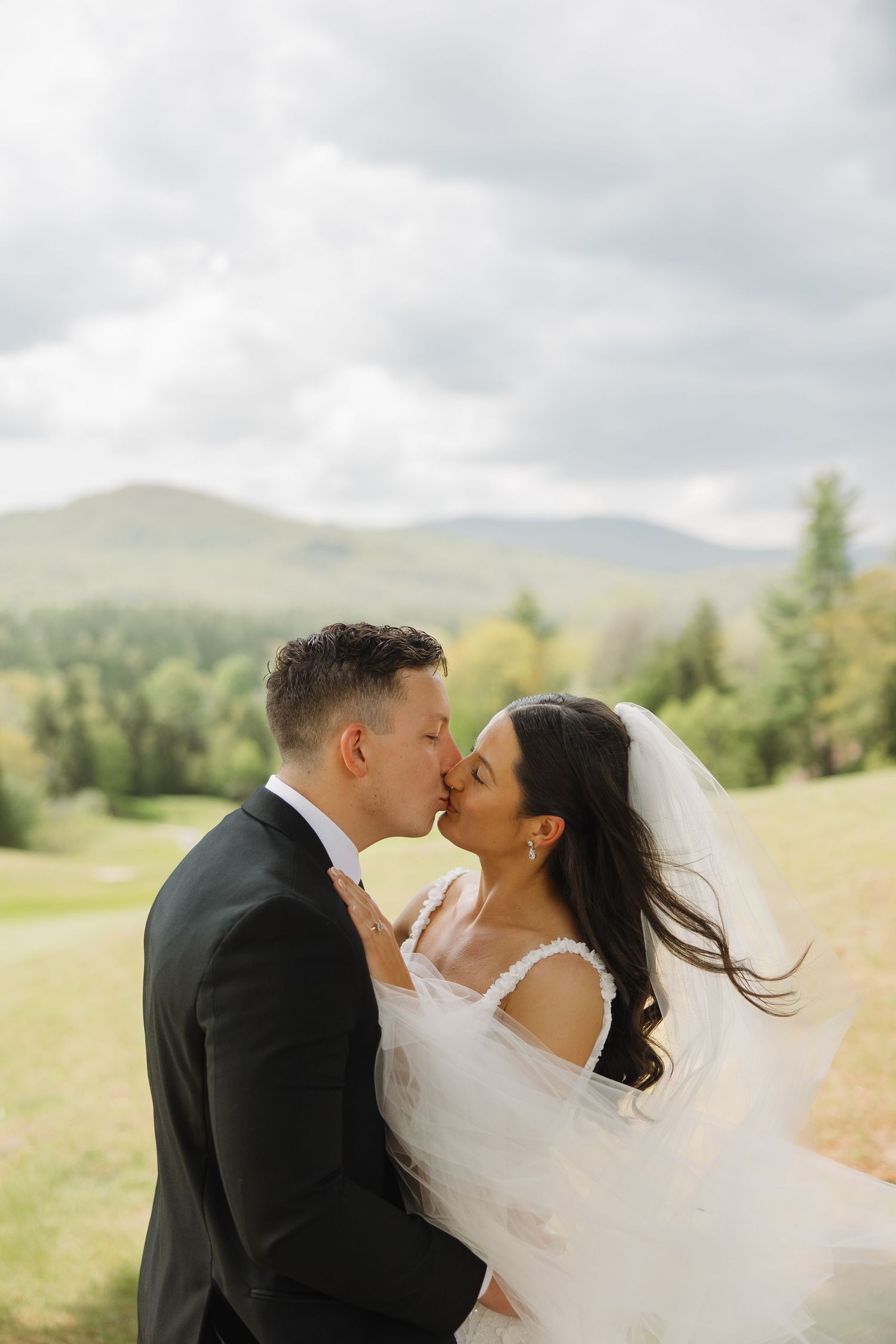 A romantic wedding couple shares an intimate kiss in a scenic mountain meadow as the bride's veil flows in the breeze.
