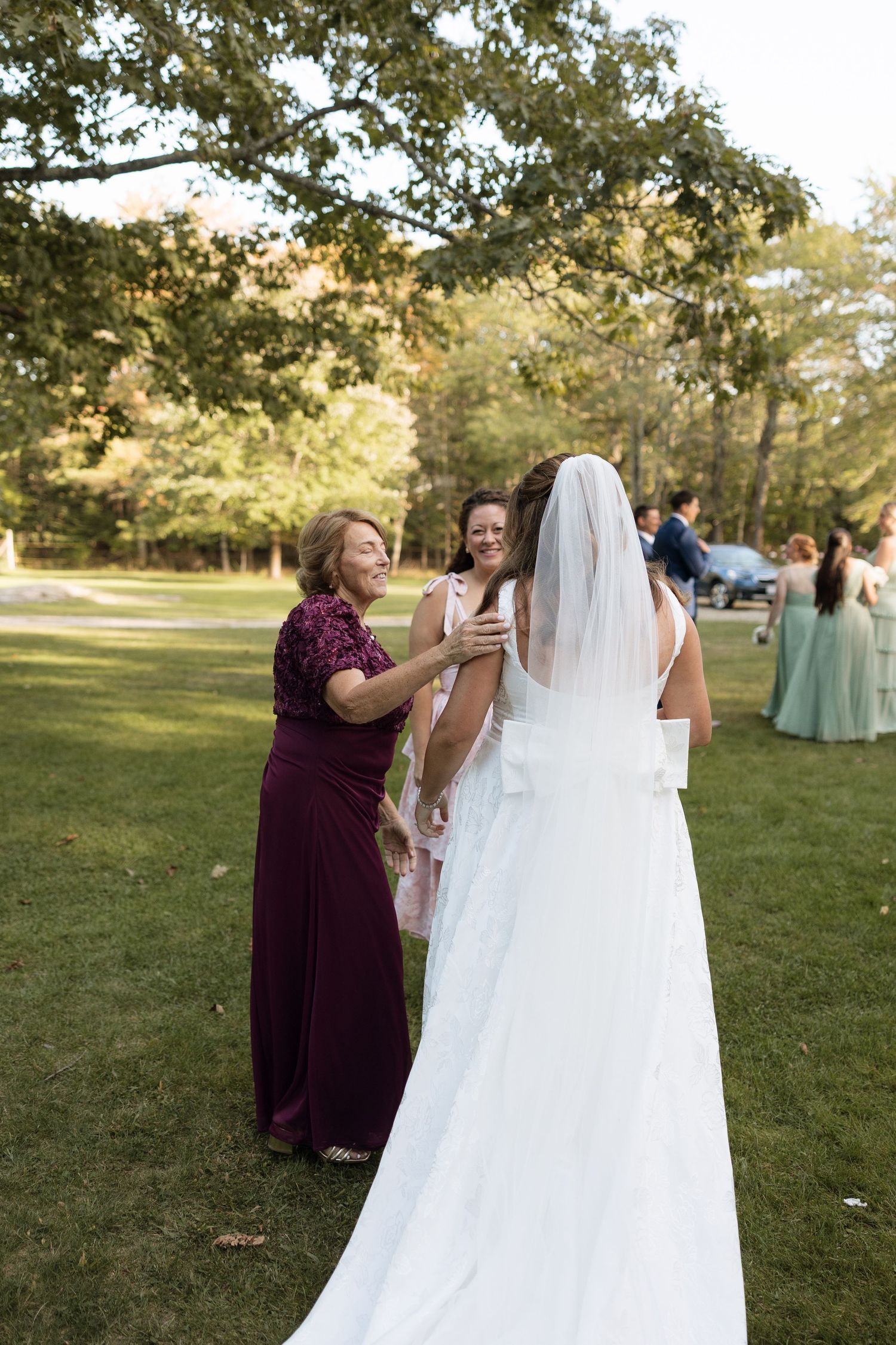 Wedding party members help adjust a flowing white veil on a bride outdoors in a sunny park setting.