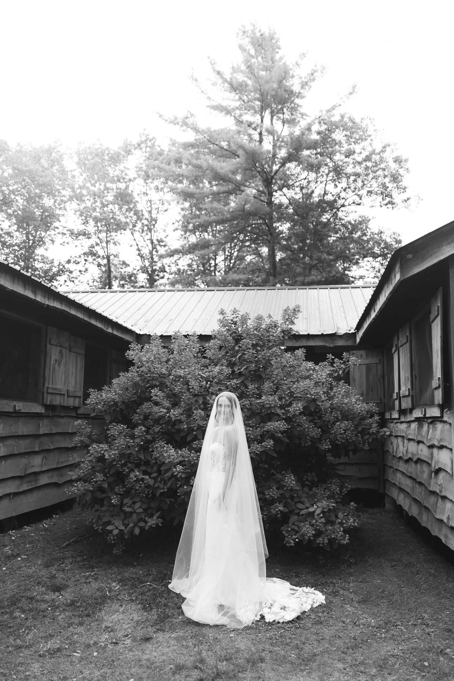 A wedding dress with a long flowing veil stands between rustic cabins surrounded by lush greenery in black and white.