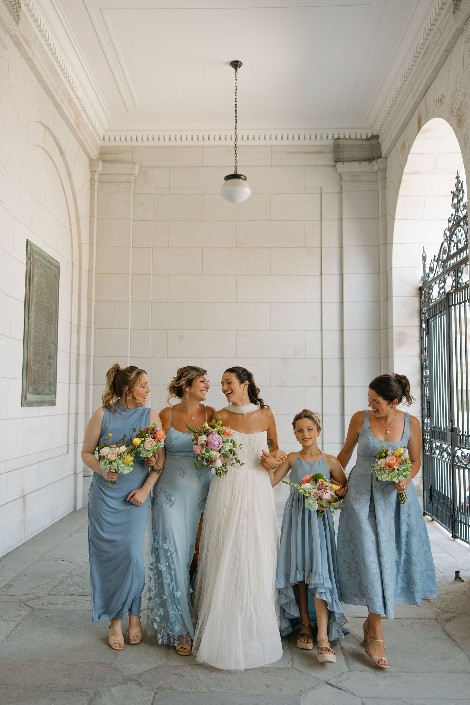 Wedding party in dusty blue bridesmaid dresses and white bridal gown share a candid moment in an elegant hallway.
