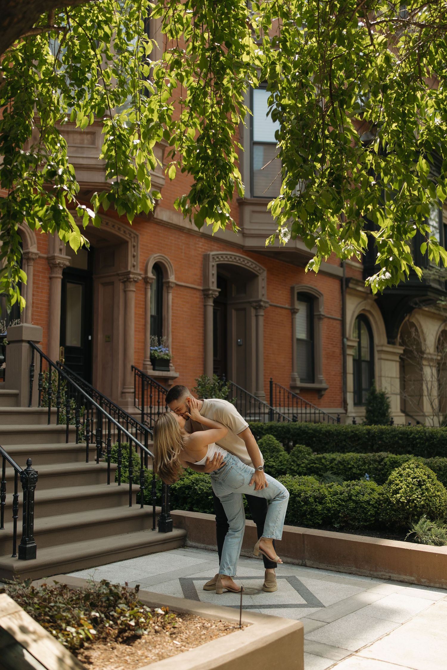 A person in casual attire performs a parkour move over a concrete planter in an urban setting with trees and brownstones.