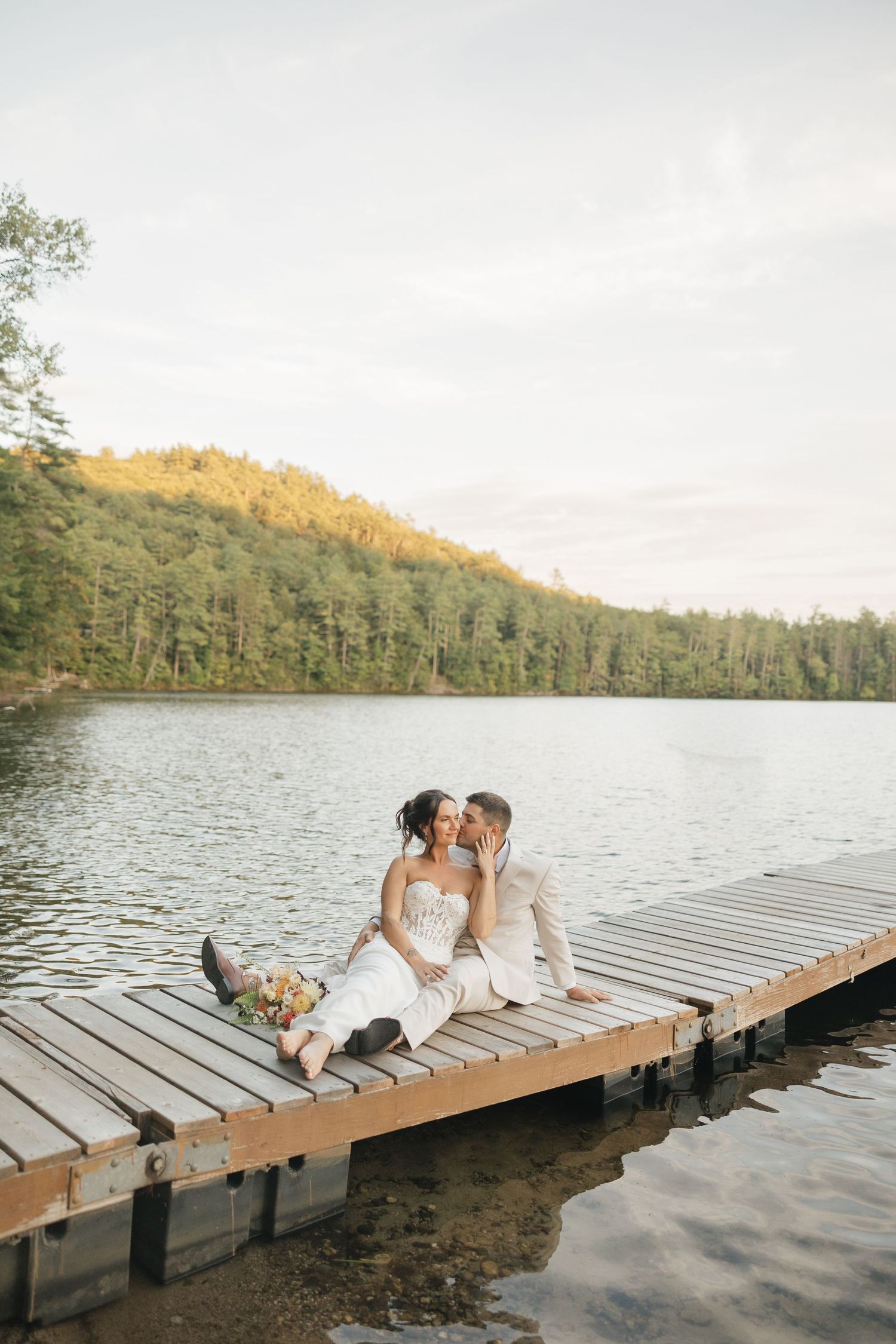 A romantic couple in white outfits share an intimate moment on a wooden dock by a serene lake at sunset.