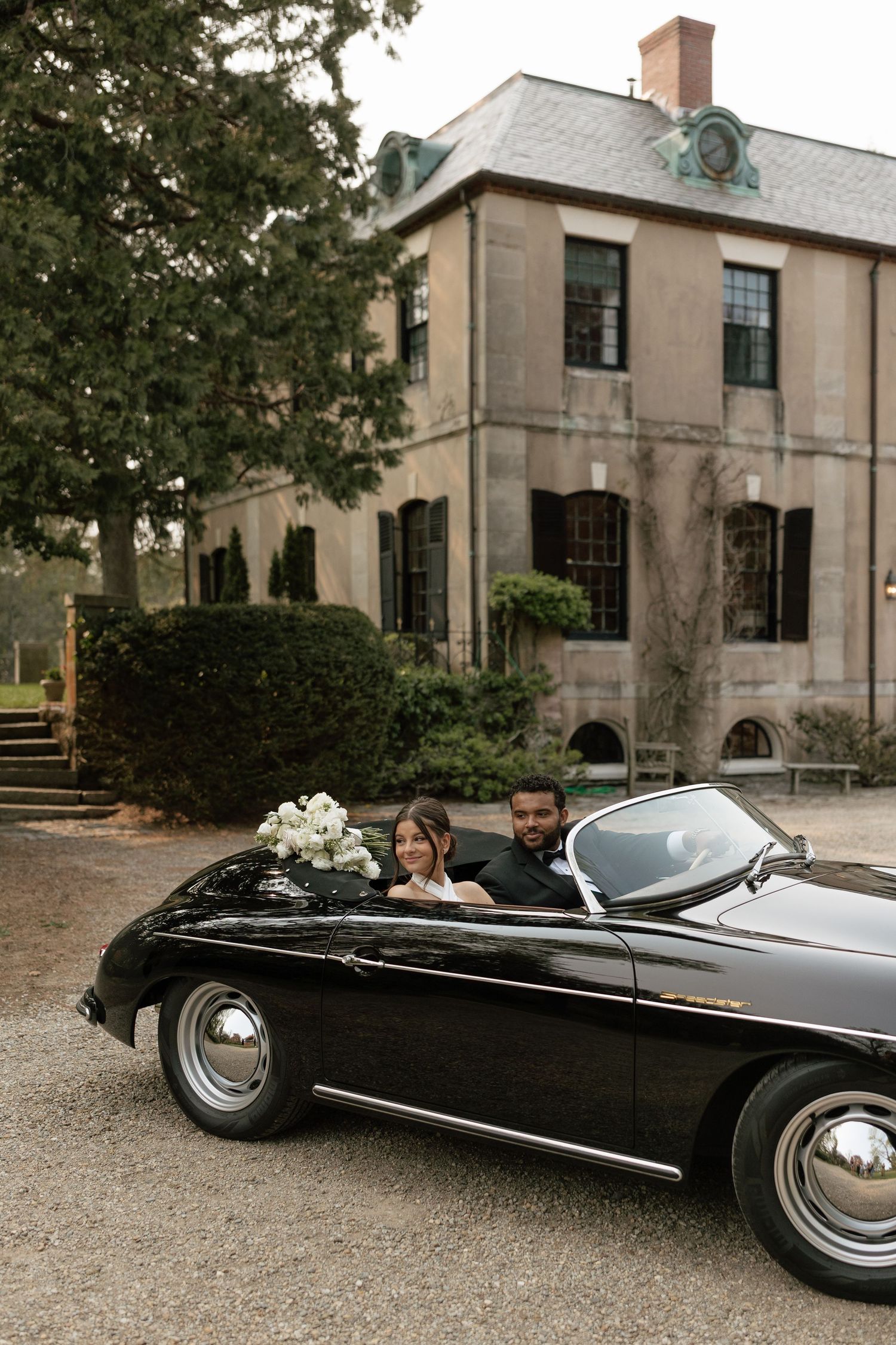 Classic black vintage Porsche convertible parked in front of an elegant stone mansion with climbing vines.