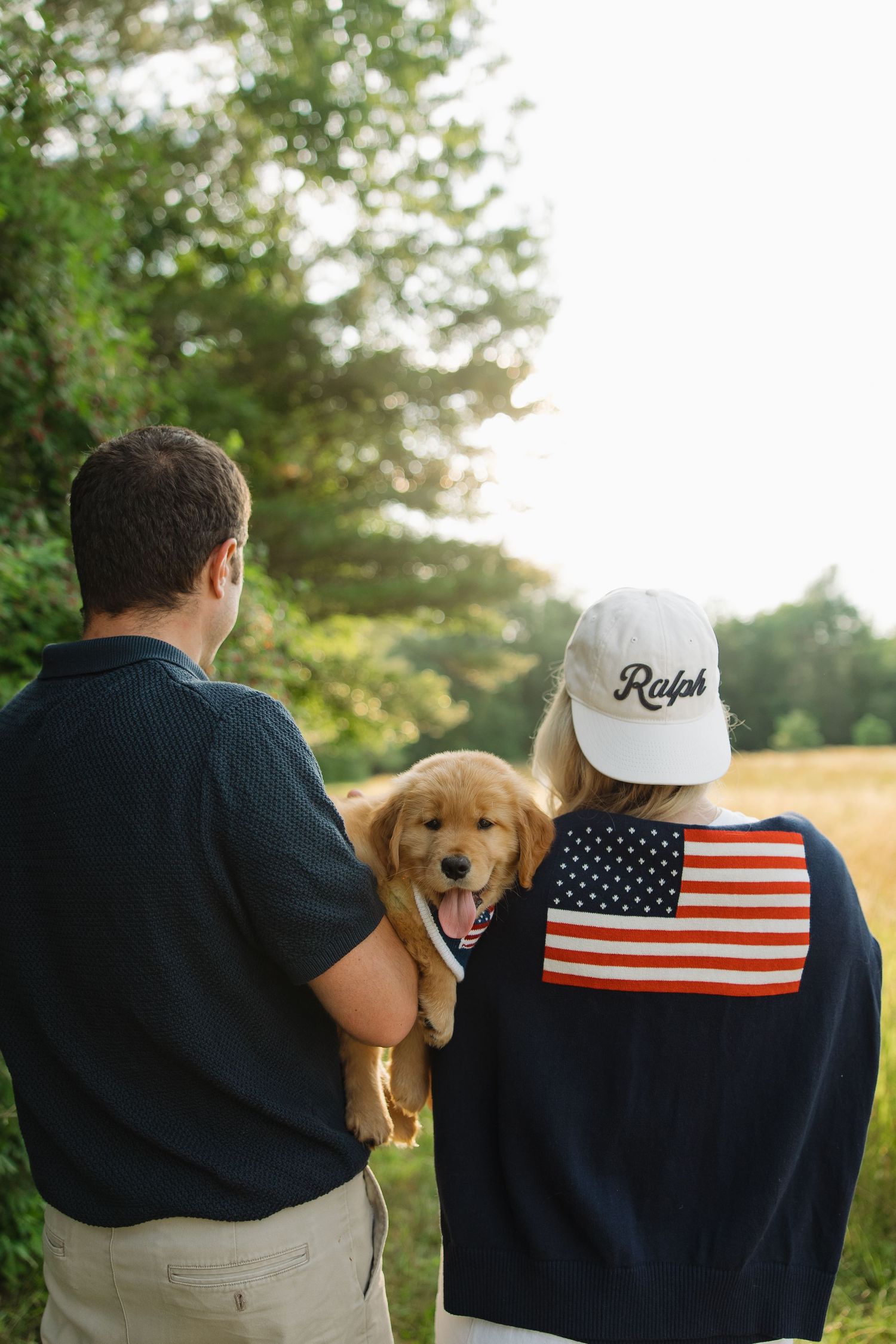 A couple sits outdoors with their golden retriever puppy while wearing patriotic American flag attire.