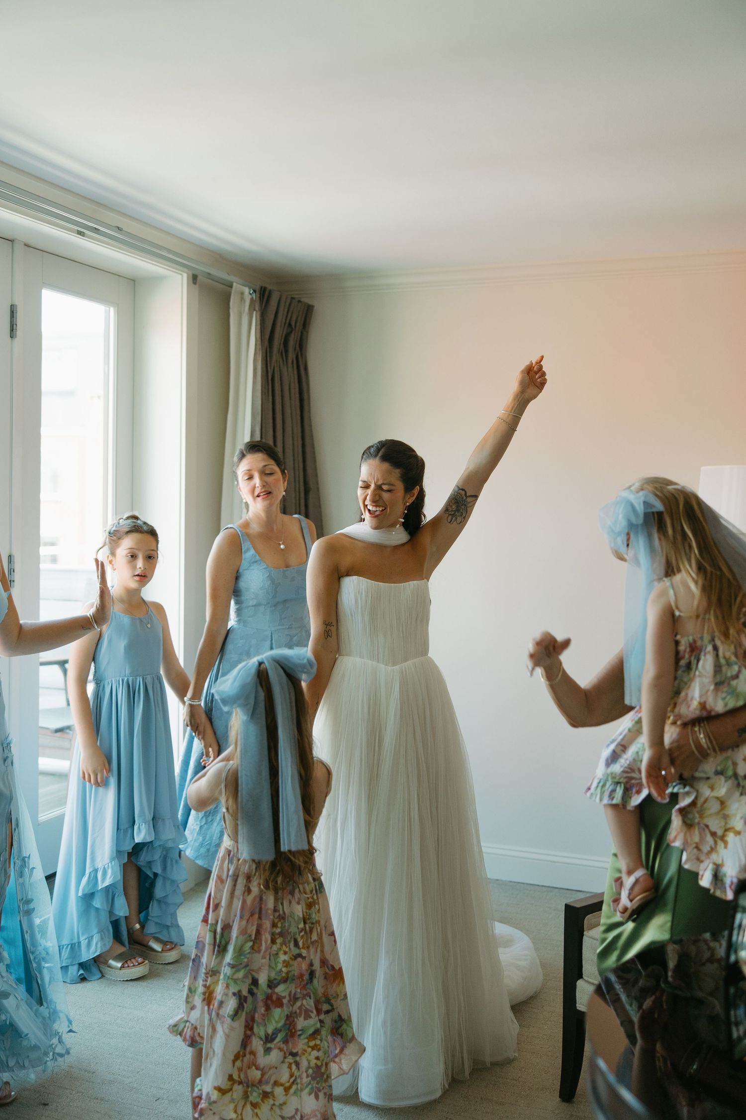 Bridesmaids in light blue dresses dance and celebrate with the bride in a white gown during wedding preparations.