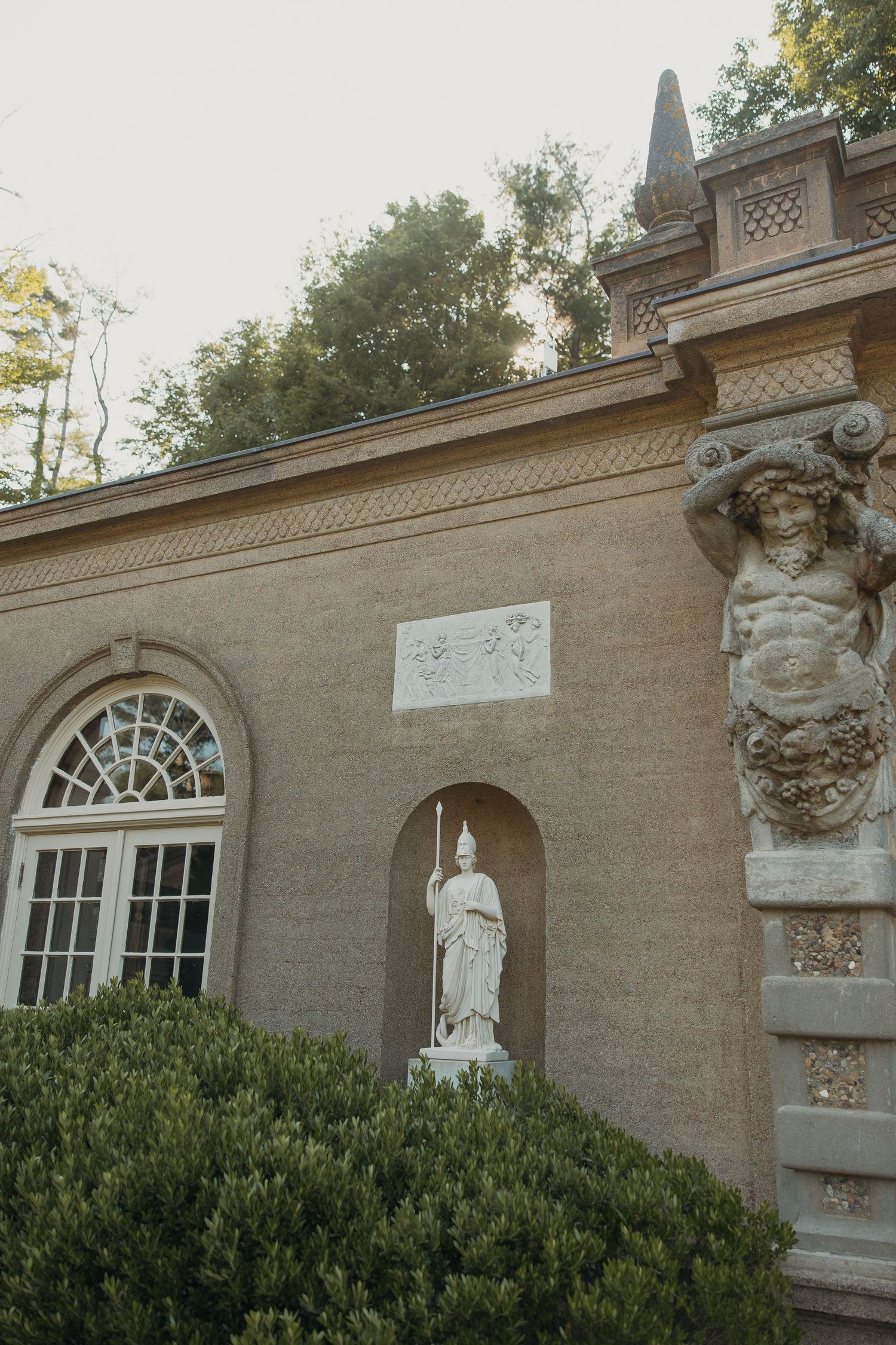 Classical stone wall architecture with arched windows and decorative statues in an elegant garden setting.