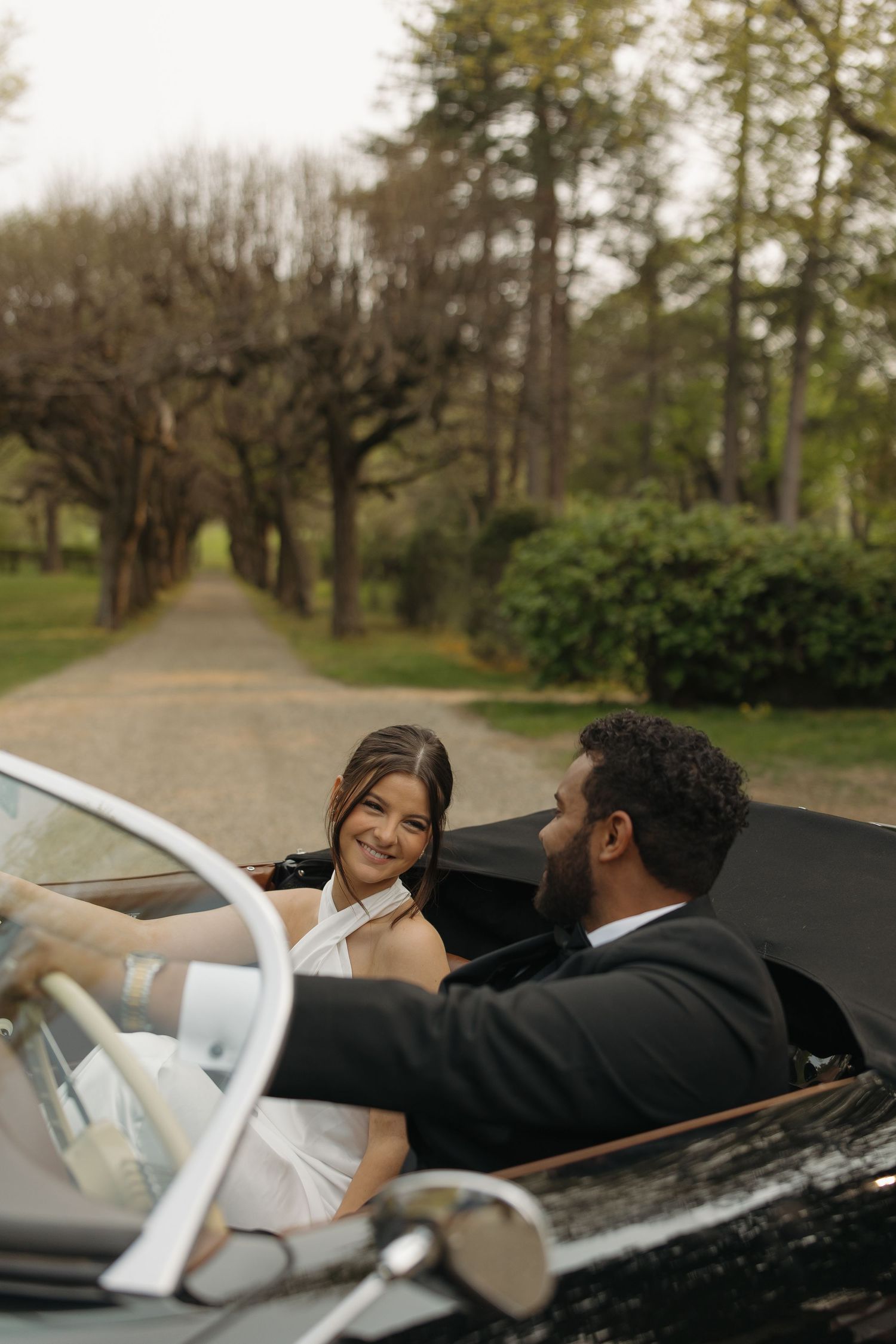 A couple in formal attire reclines in a vintage convertible car on a tree-lined path.