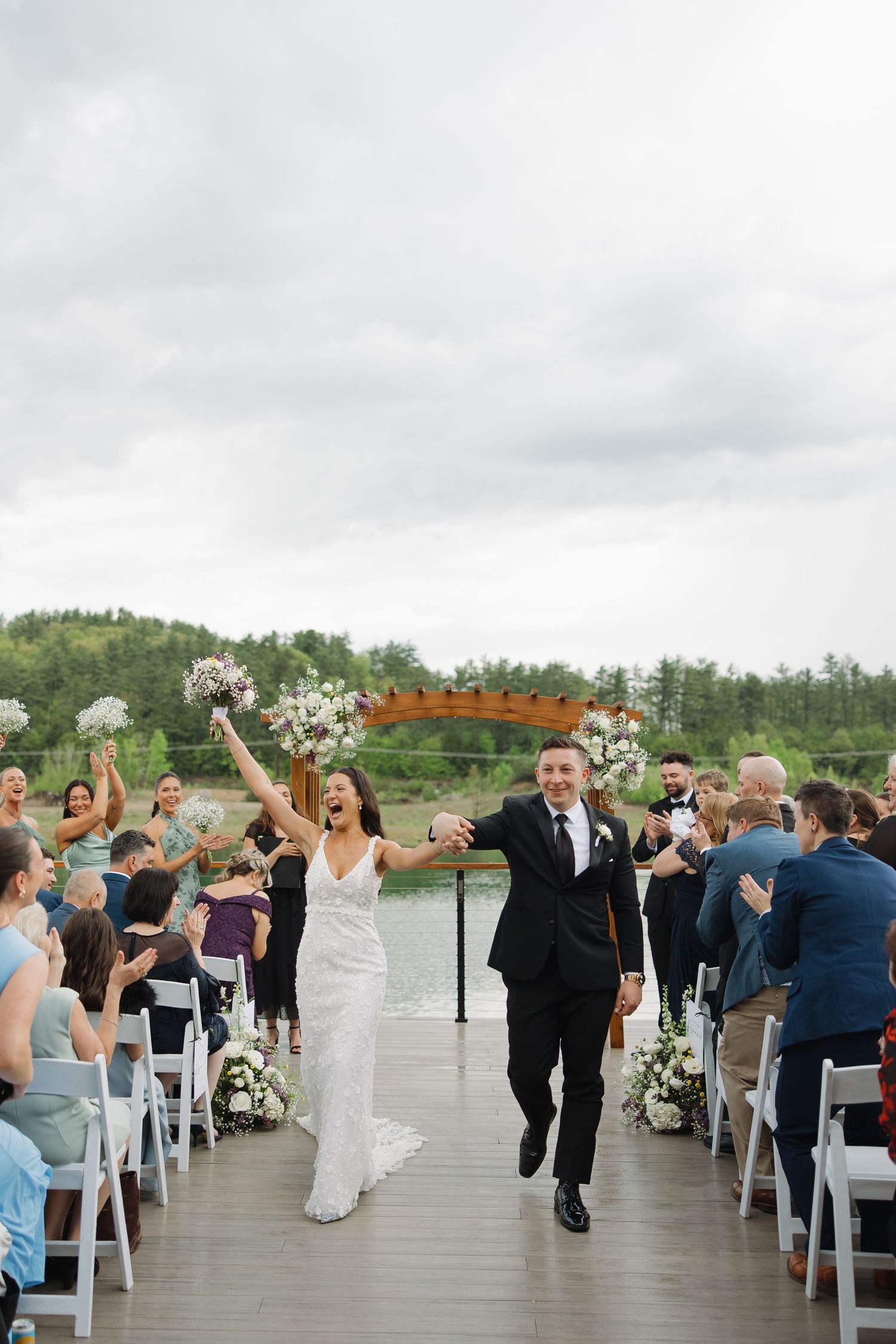 Wedding guests celebrate and dance on an outdoor terrace overlooking a scenic lake and forest landscape.