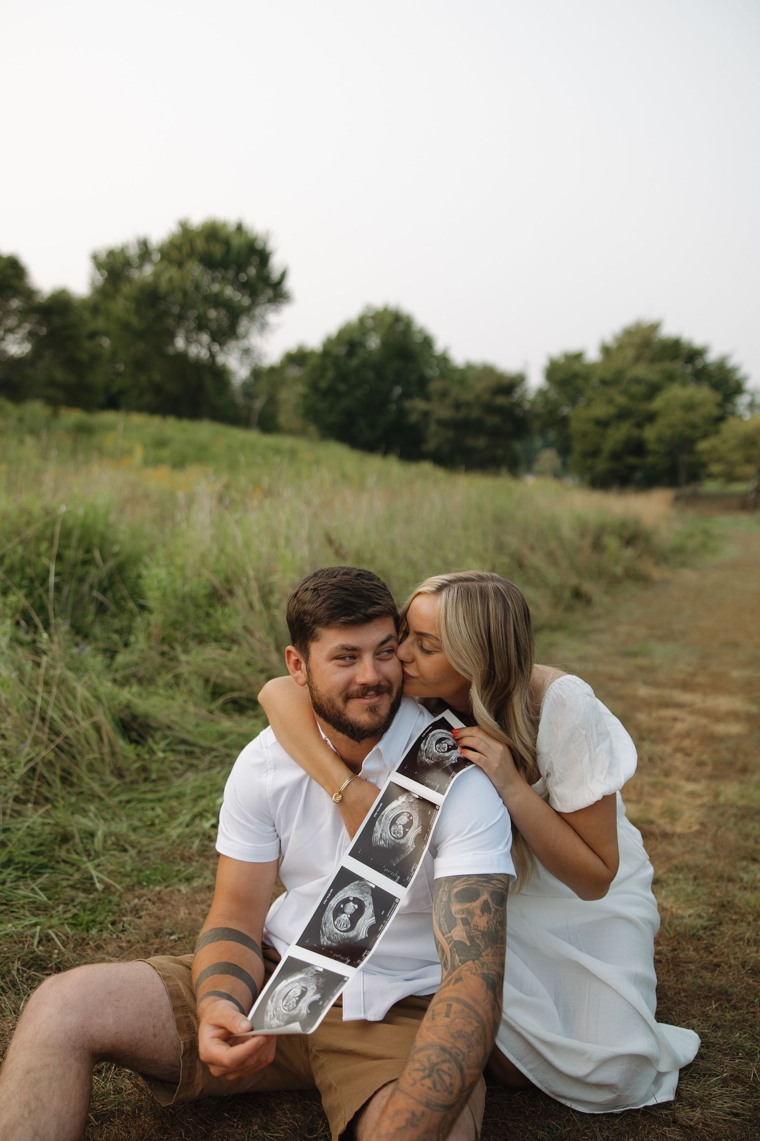 A couple sits in a grassy field sharing ultrasound photos and tender moments together.