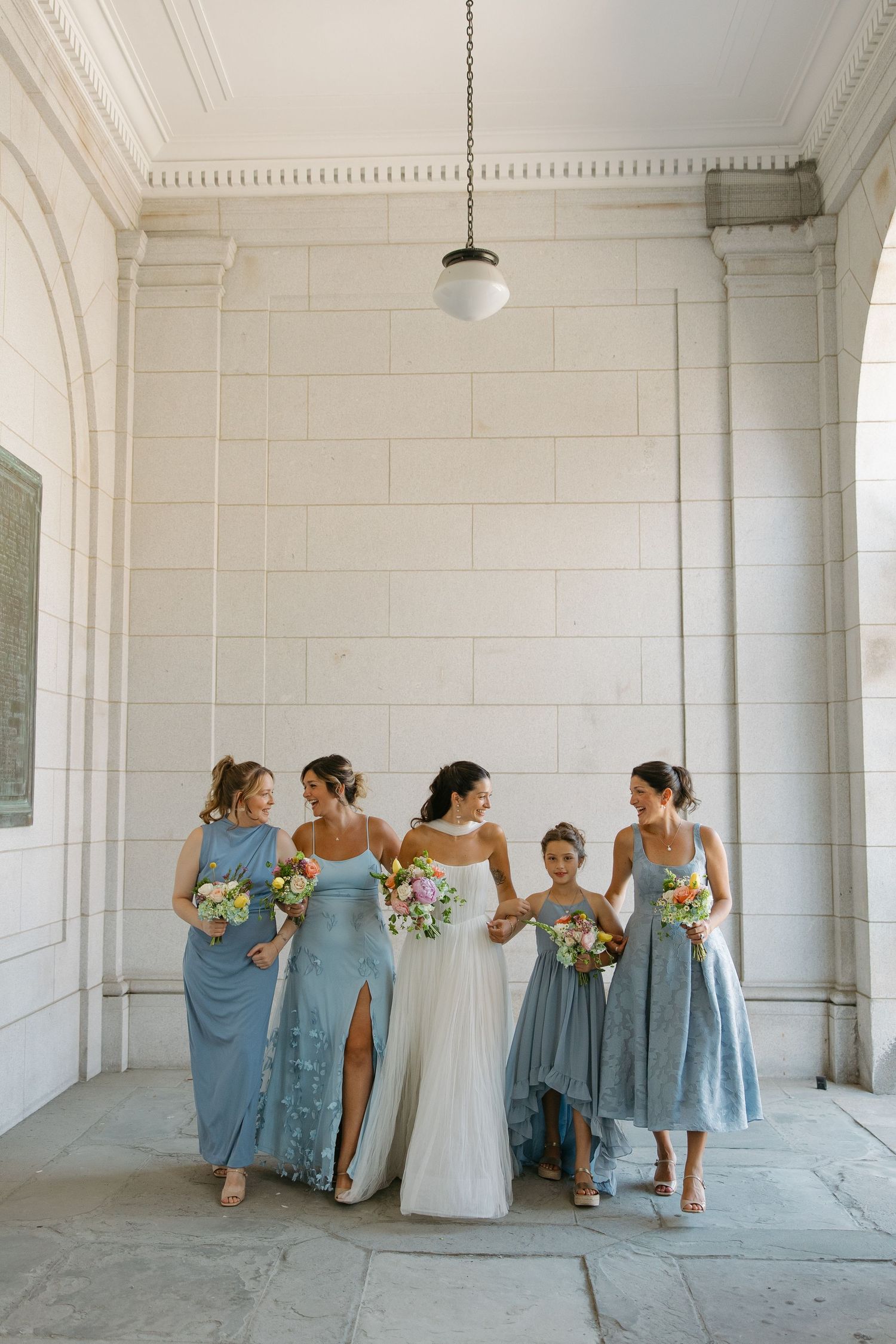 Bridesmaids in dusty blue dresses share candid moments with the bride in a bright white hallway setting.