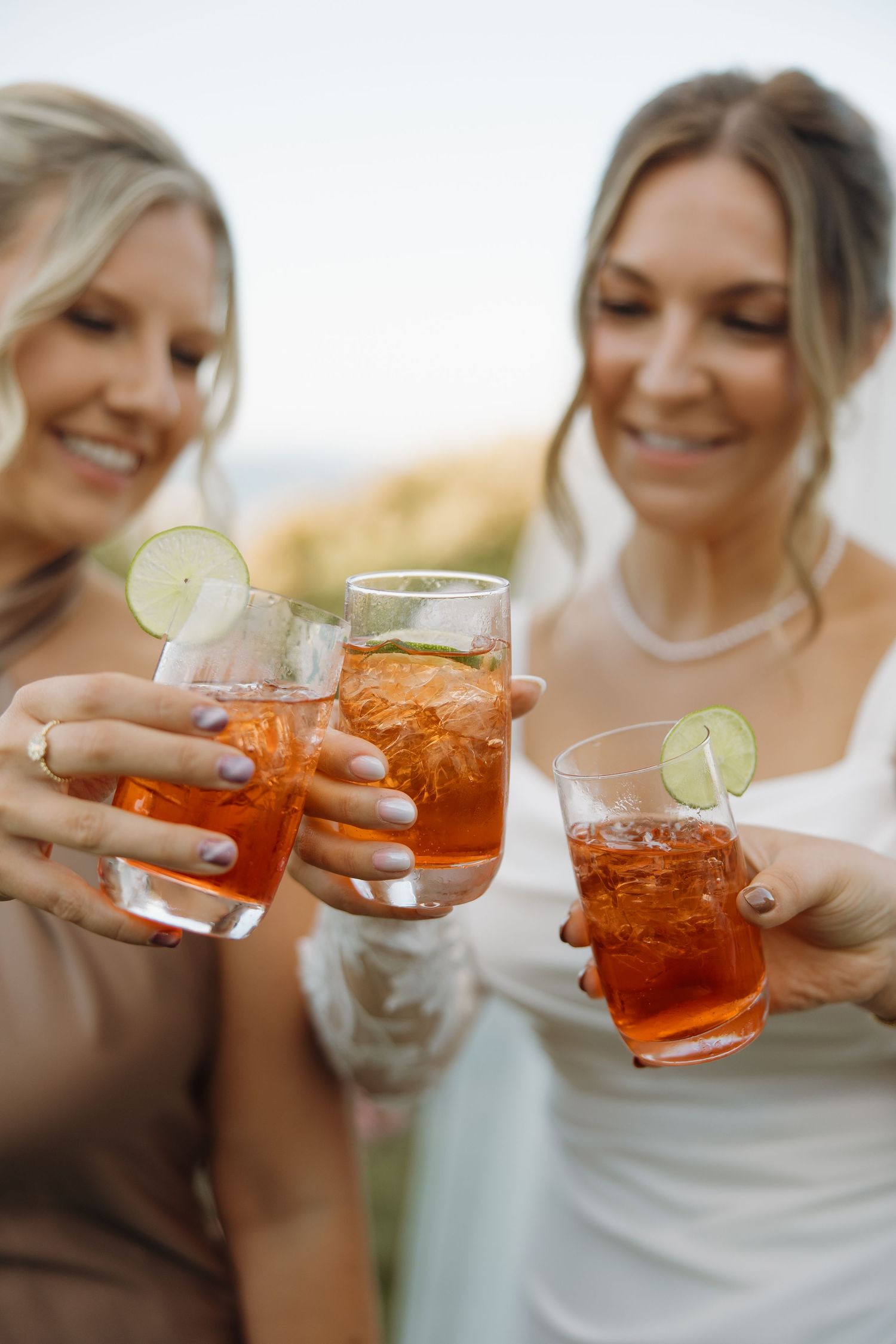 Friends in white dresses clink glasses filled with orange Aperol Spritz cocktails during a celebration.