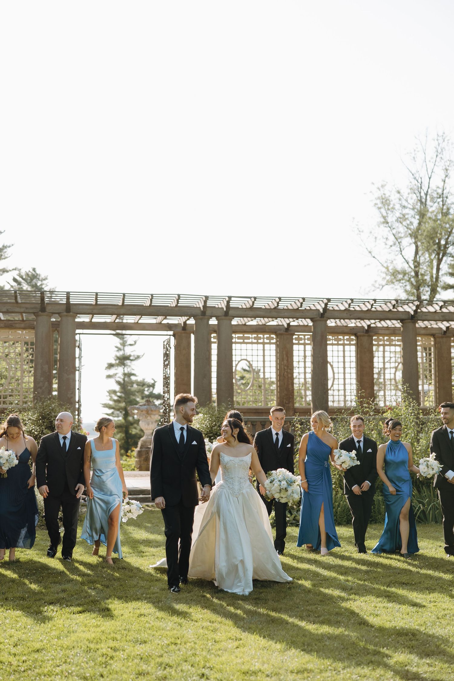 Wedding party walks on grass under a pergola on a sunny day with bridesmaids in blue dresses.