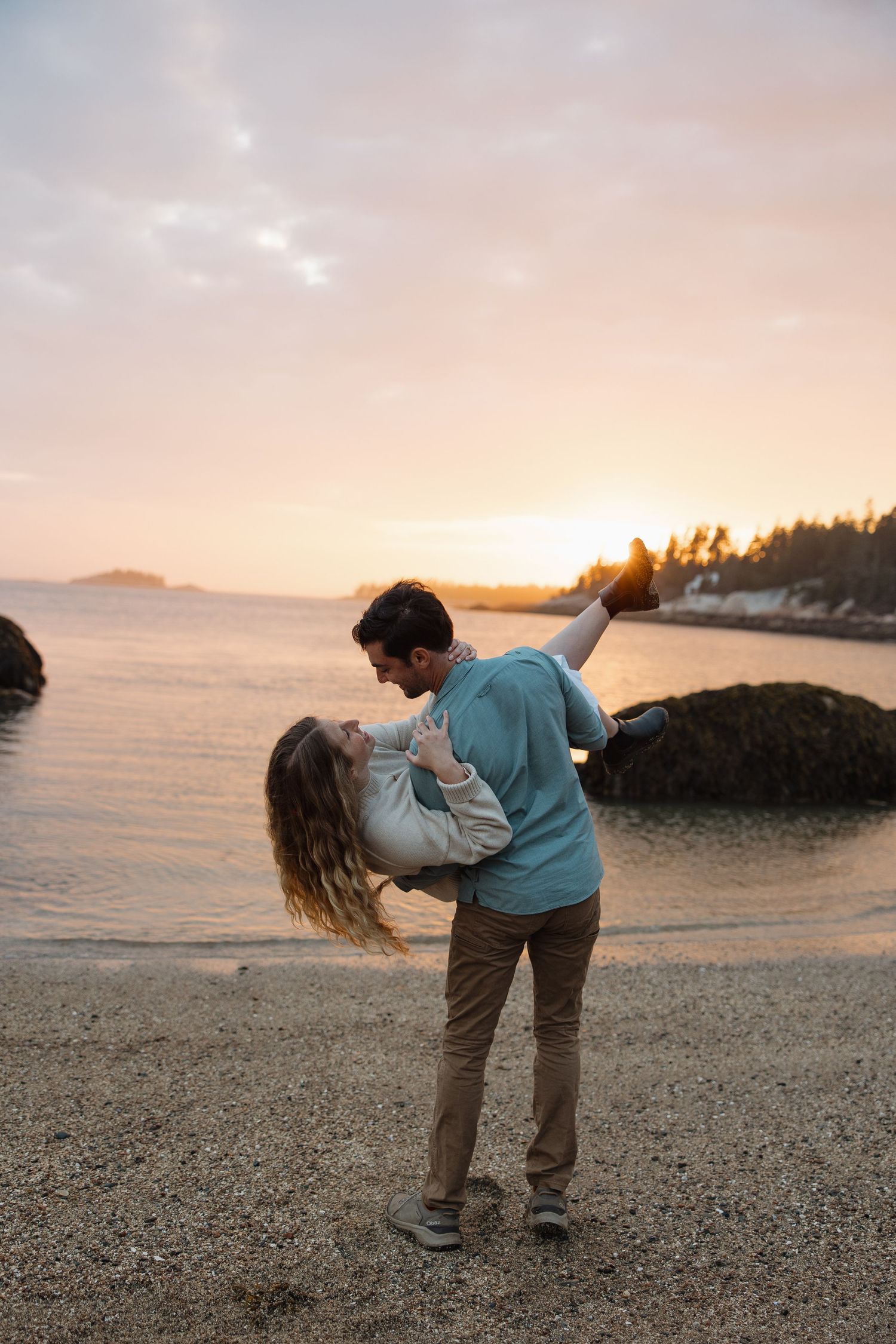 A romantic couple dances on a beach at sunset with the ocean and coastline in the background.