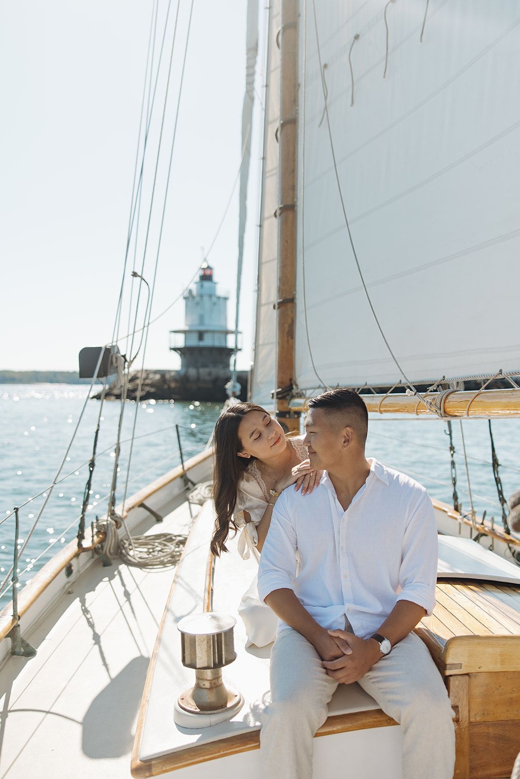 A romantic couple in white outfits sharing intimate moments aboard a sailboat with a lighthouse visible in the background.
