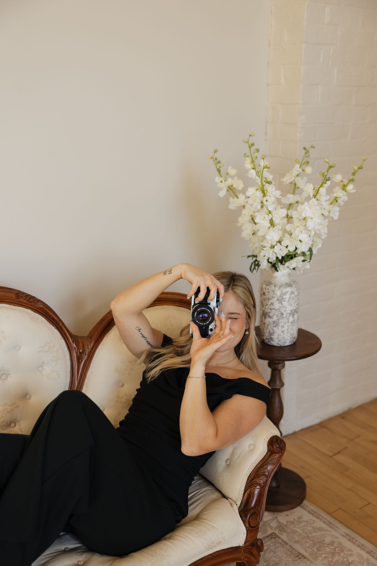 A person in black dress reclines on a vintage sofa with white flowers in a vase nearby.